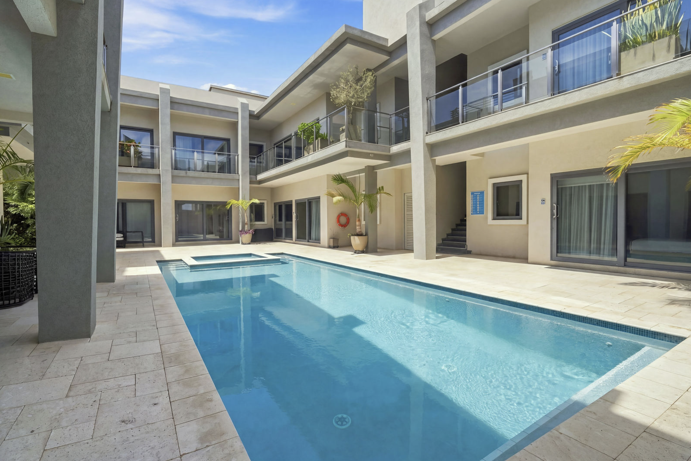The image shows a modern apartment complex with a swimming pool in the courtyard. The buildings have multiple stories with balconies and sliding glass doors. There are potted plants and palm trees around the pool area, and the sky is clear with a few