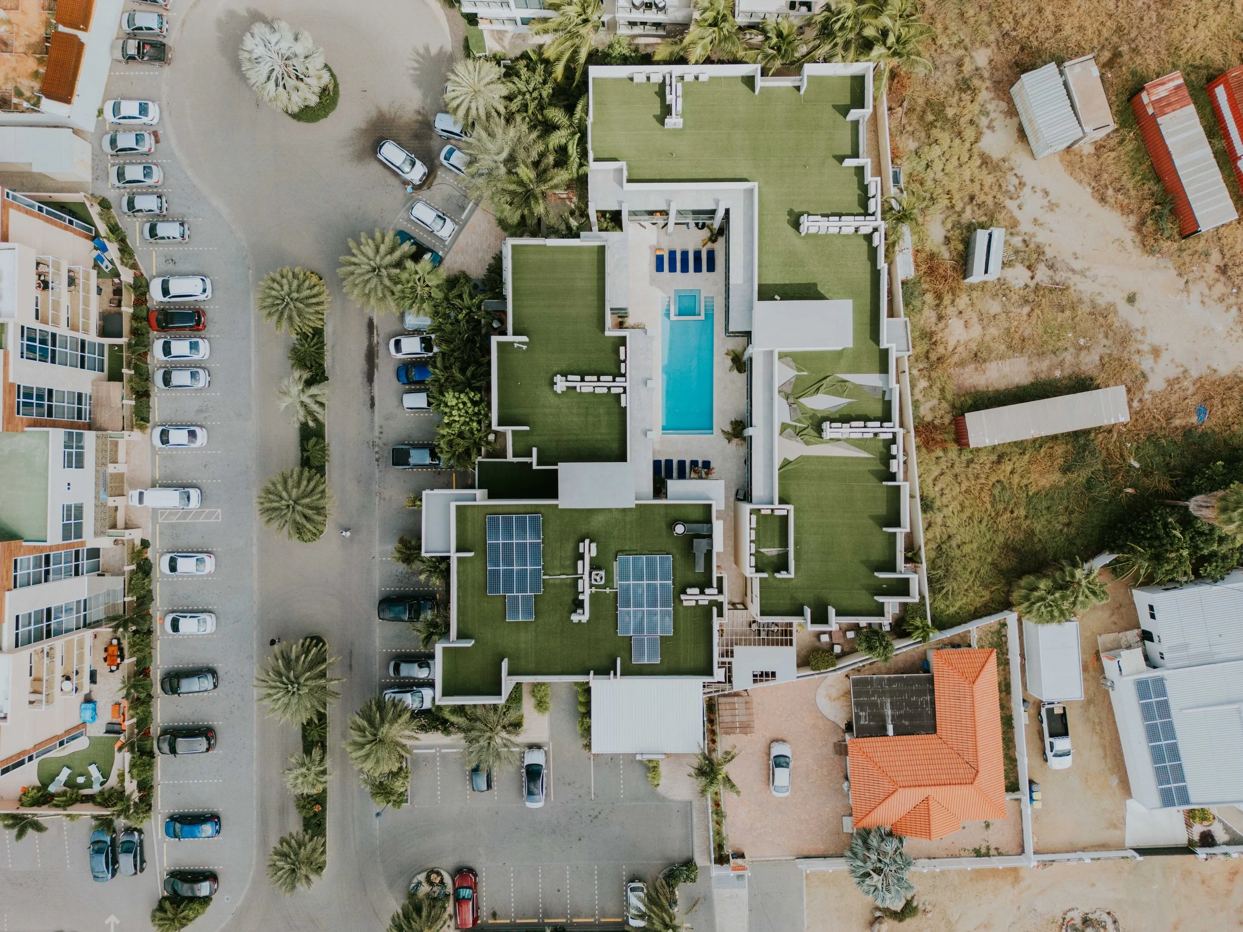 An aerial view of a building with green roof sections, a swimming pool, and solar panels, surrounded by parking lots, trees, and neighboring structures.