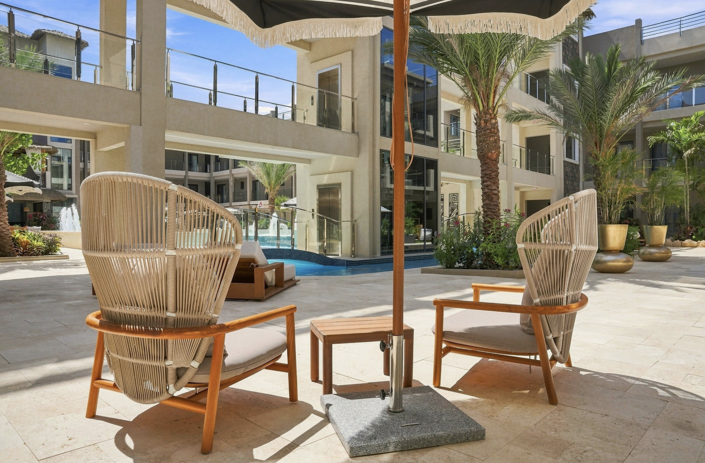 Outdoor patio area with wicker chairs, a large beige umbrella, palm trees, potted plants, and a building with balconies in the background.