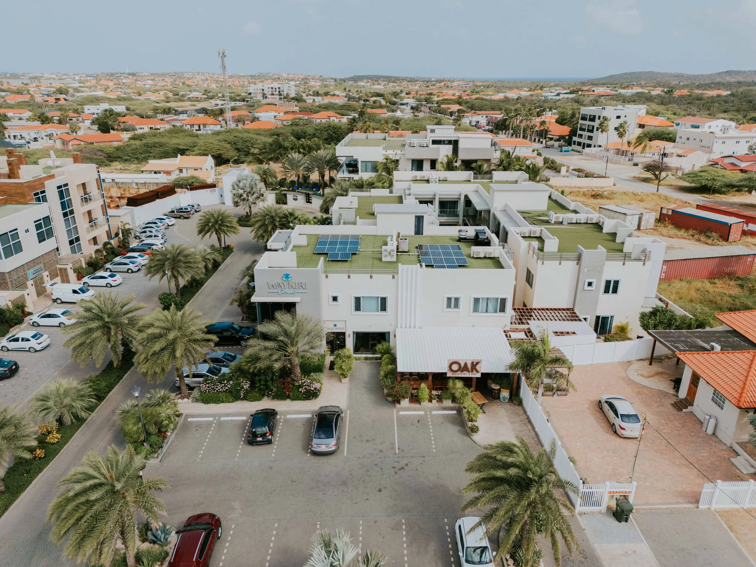 An aerial view of a modern commercial and residential area with a building labeled 'OAK' and another labeled 'WAYKIRI'. The scene includes parking lots, palm trees, and nearby houses with red-tiled roofs.