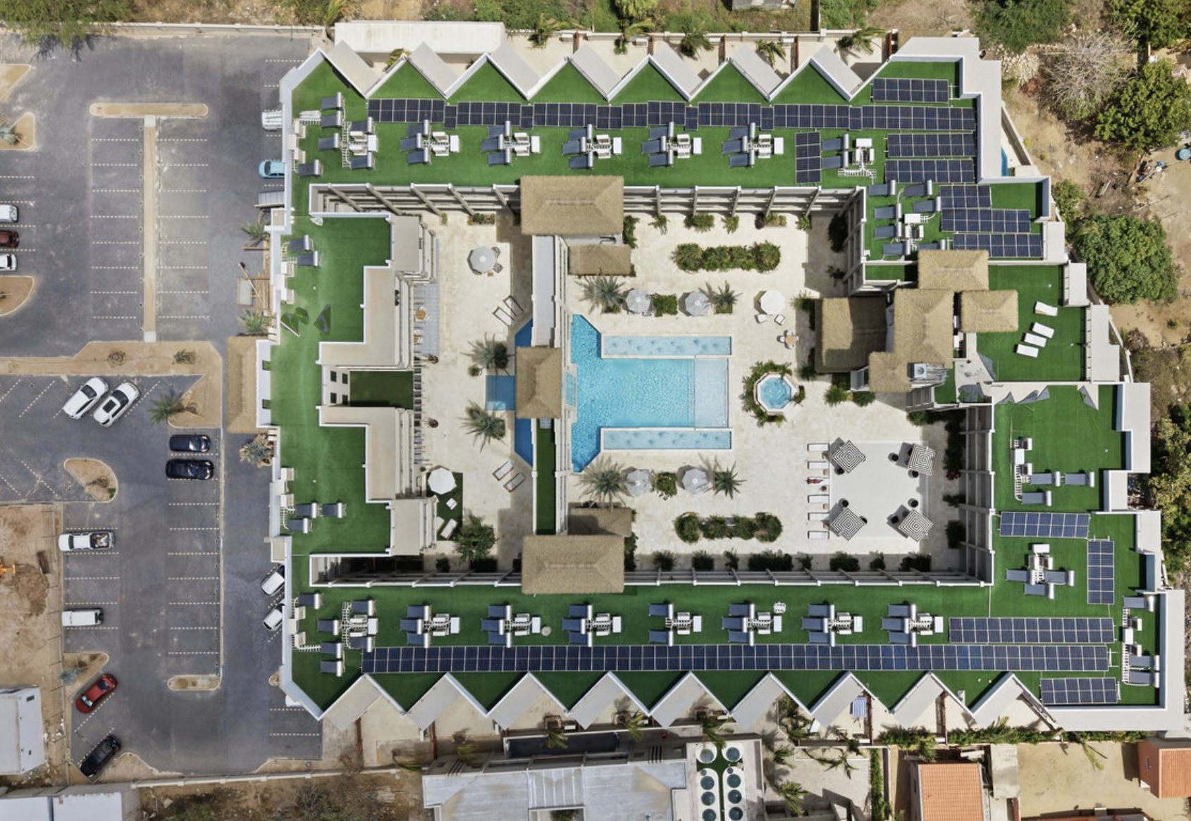 Aerial view of a large residential building with multiple green rooftops, solar panels, a central swimming pool, and surrounding greenery. Adjacent parking lot with cars on the left side.