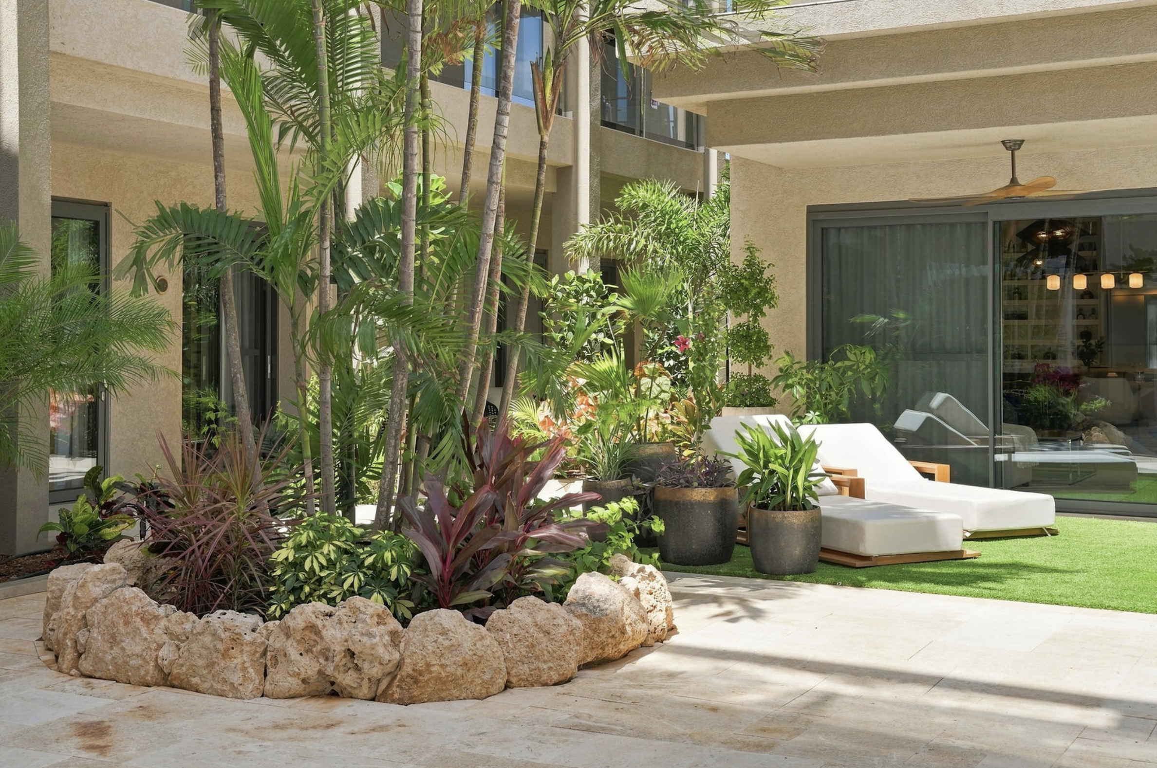 A lush courtyard with various tropical plants and trees, modern beige apartment building in the background, outdoor seating with white lounge chairs, sliding glass doors, and a ceiling fan.