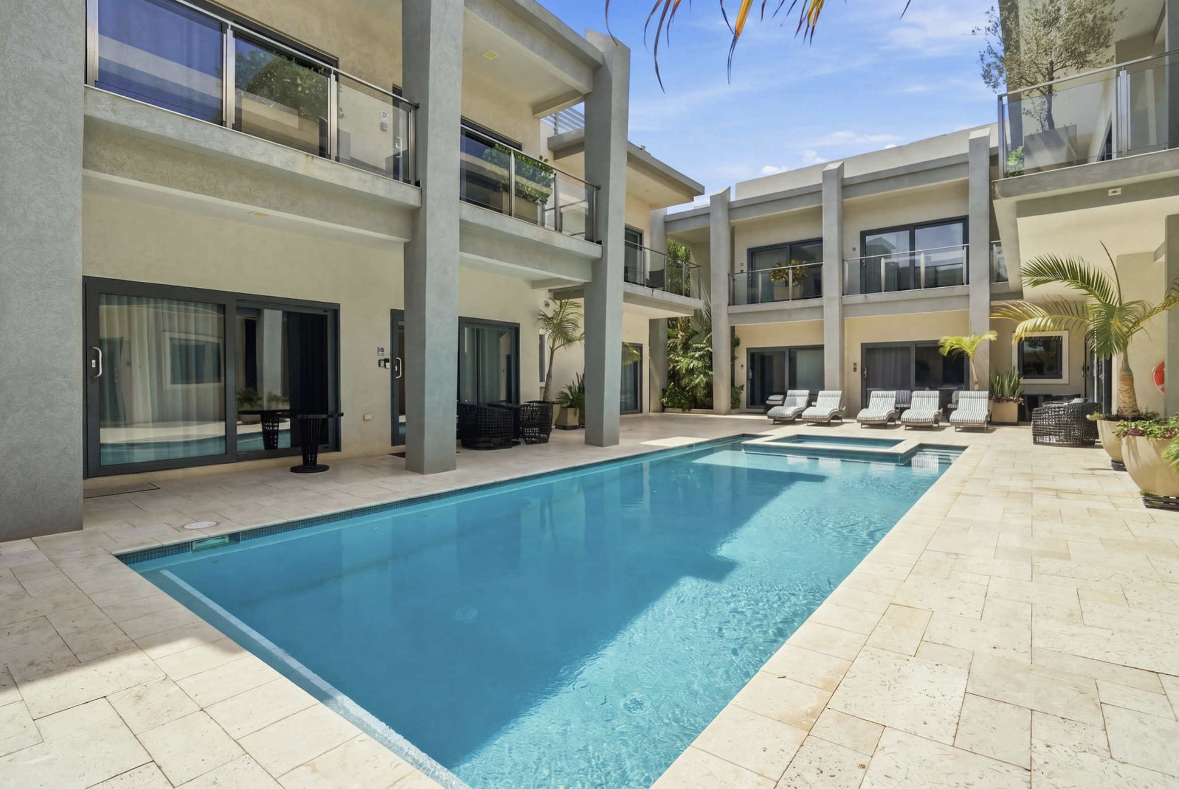 A modern apartment complex with a swimming pool, lounge chairs, and outdoor seating area surrounded by palm plants.