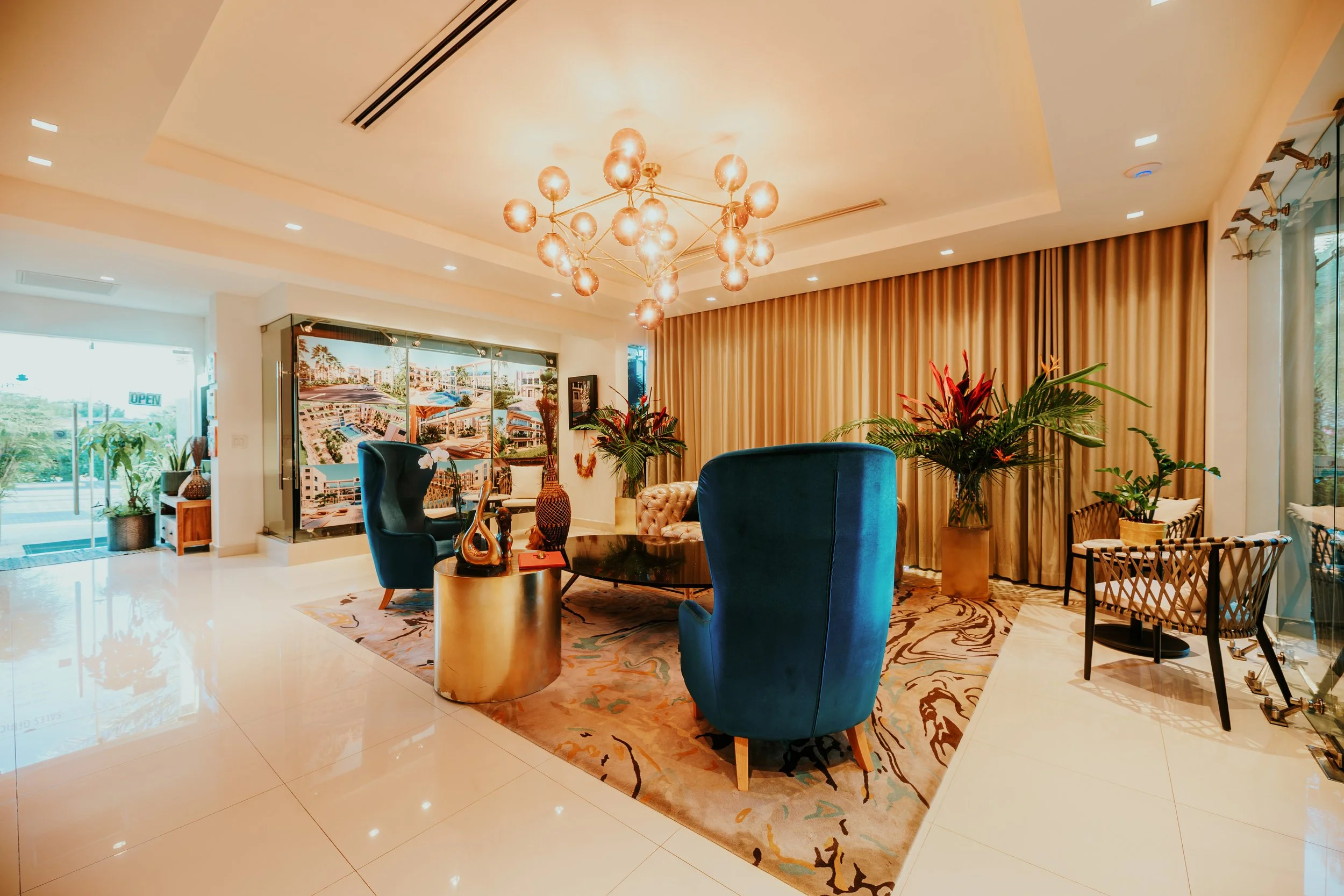 Modern hotel lobby with blue armchairs, a black glass coffee table, large potted plants, a colorful rug, and a golden chandelier.