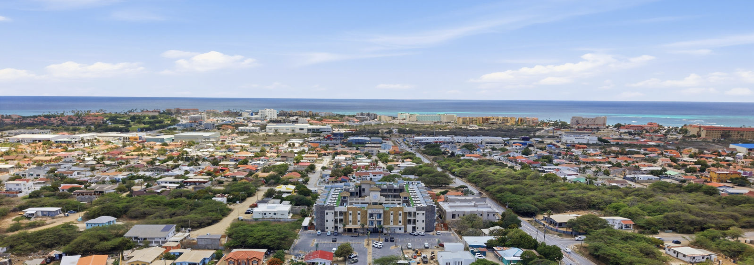 Aerial view of a coastal city with colorful buildings, greenery, and a beach with ocean in the background on a partly cloudy day.