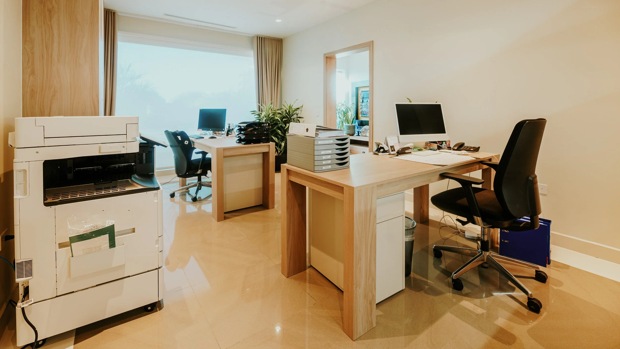 Office space with two desks, computer monitors, office chairs, a printer, and filing trays, illuminated by natural light from a large window with beige curtains.