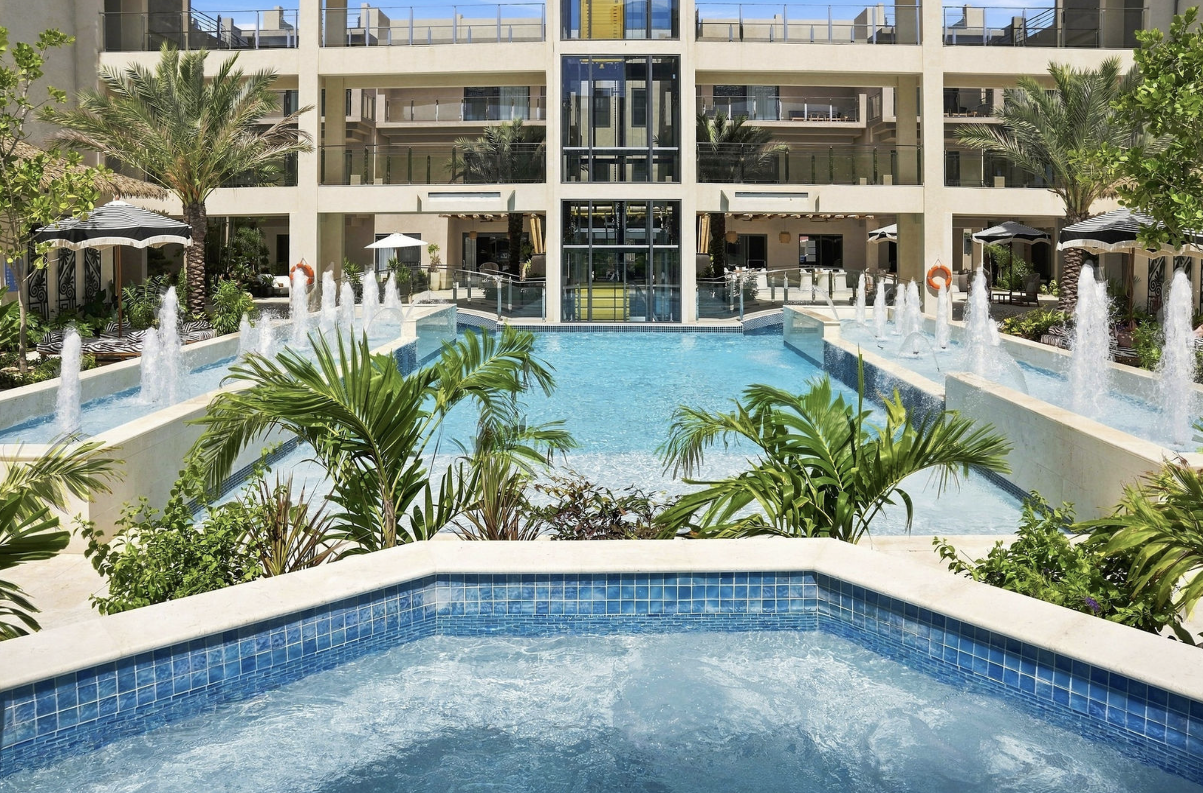 Exterior view of a hotel with multiple balconies, pool area with fountains, palm trees, umbrellas, and lounge chairs.