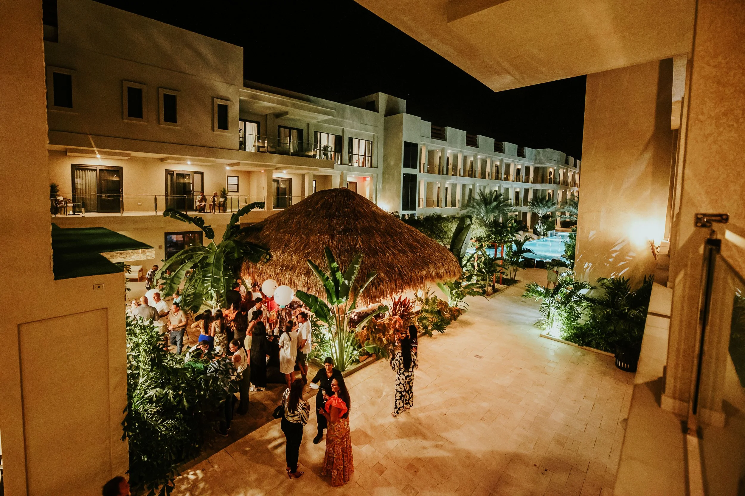 Nighttime scene at a hotel pool area with a thatched-roof cabana, tropical plants, and an outdoor pool. People are gathered and socializing near the cabana, illuminated by warm lighting.