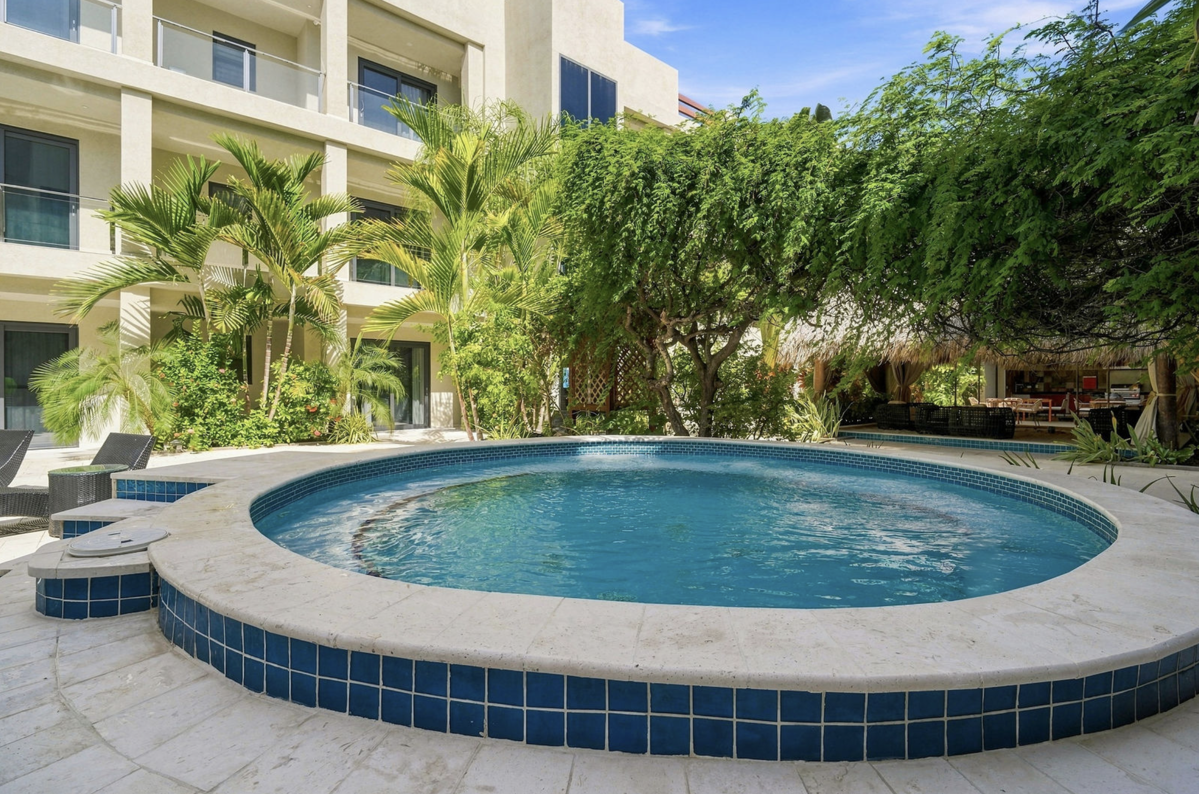 A backyard scene with a swimming pool surrounded by a white tiled deck, lush green tropical plants including palm trees, and a multi-story light-colored apartment building with balconies in the background. The sky is clear and blue.