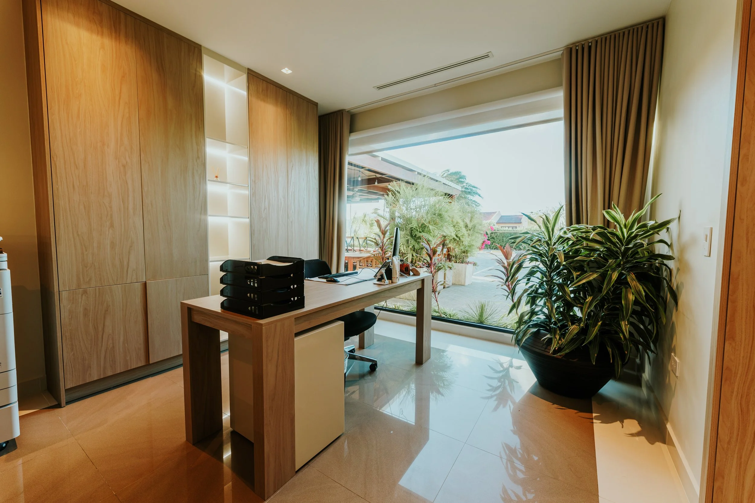 Modern office with wooden furniture and large glass window with outdoor view and plants.