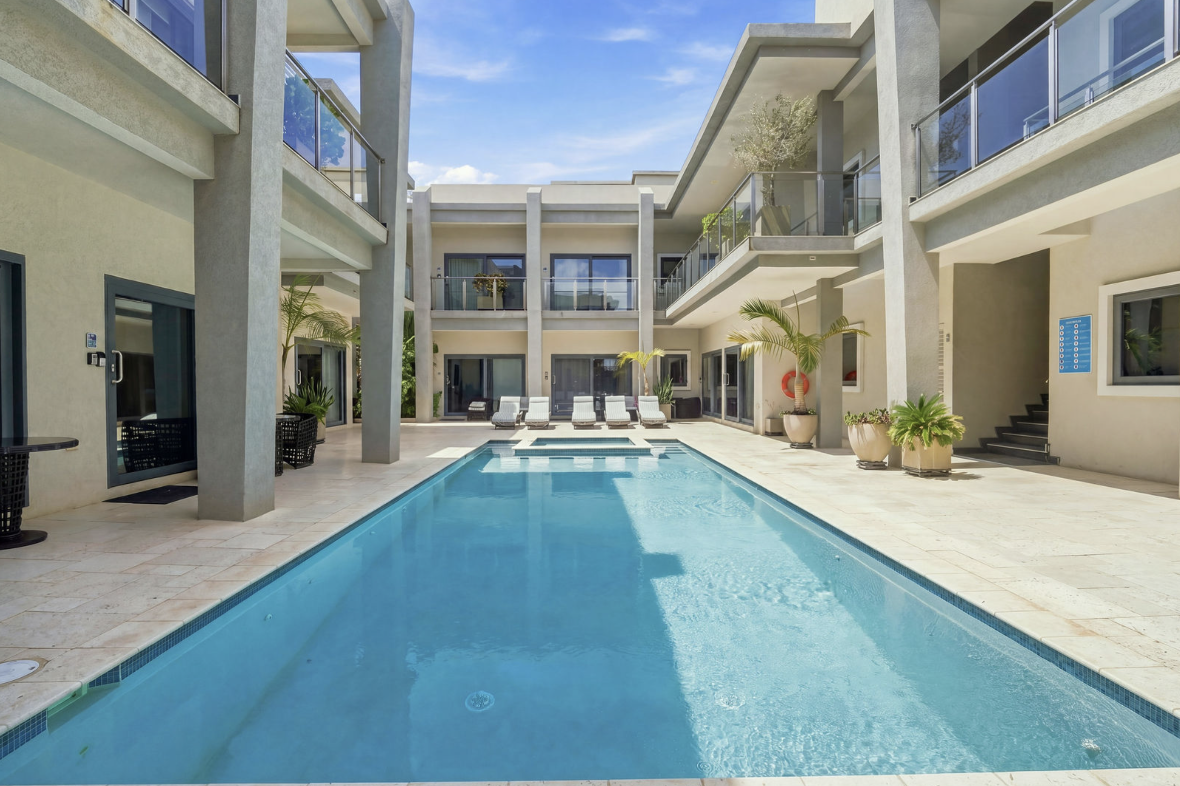 Modern apartment courtyard with a swimming pool, lounge chairs, and potted plants, surrounded by a multi-story building with balconies and large windows.