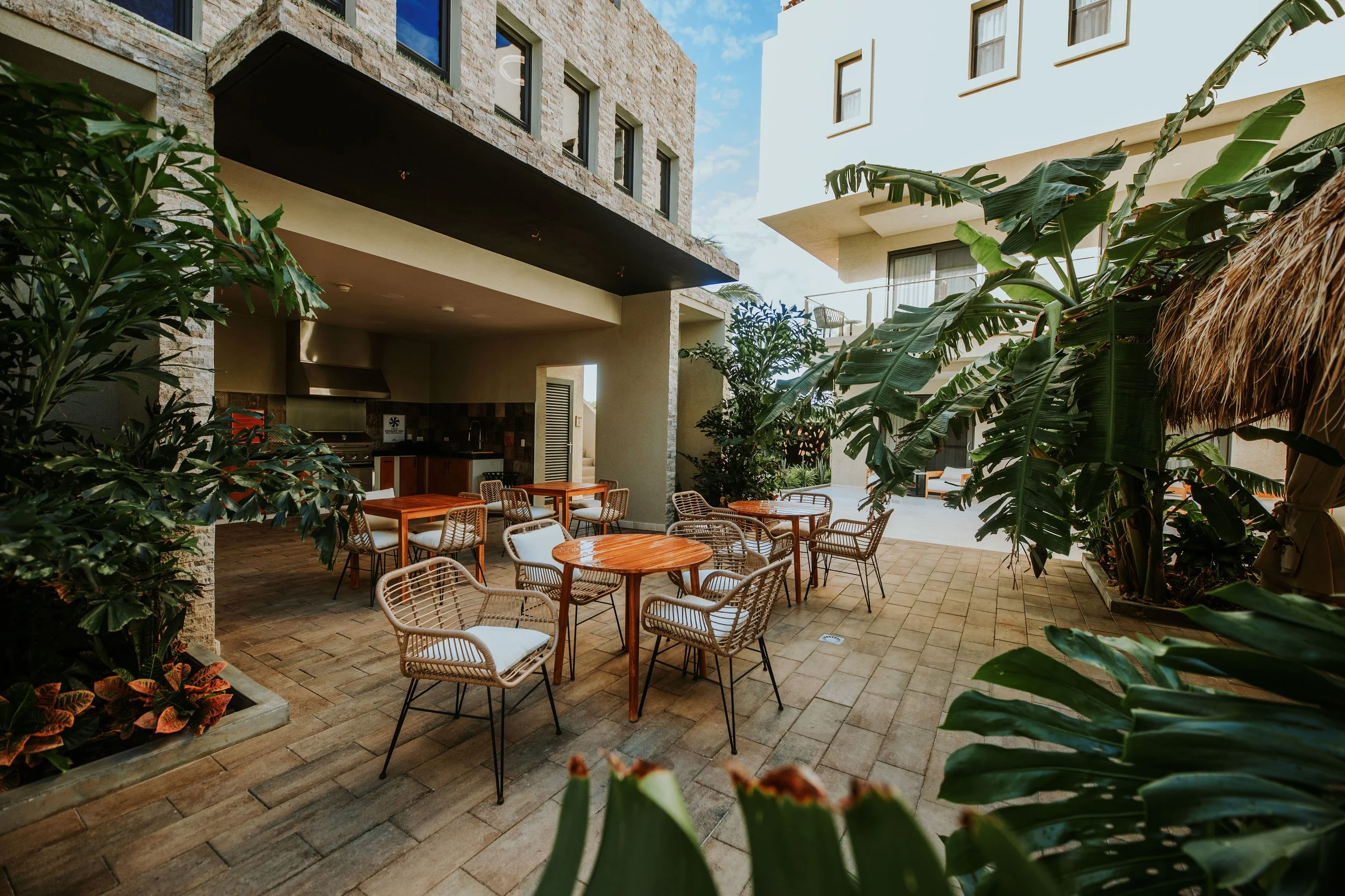 An outdoor patio area with wooden tables and wicker chairs, surrounded by tropical plants, with a building in the background, under a partly cloudy sky.