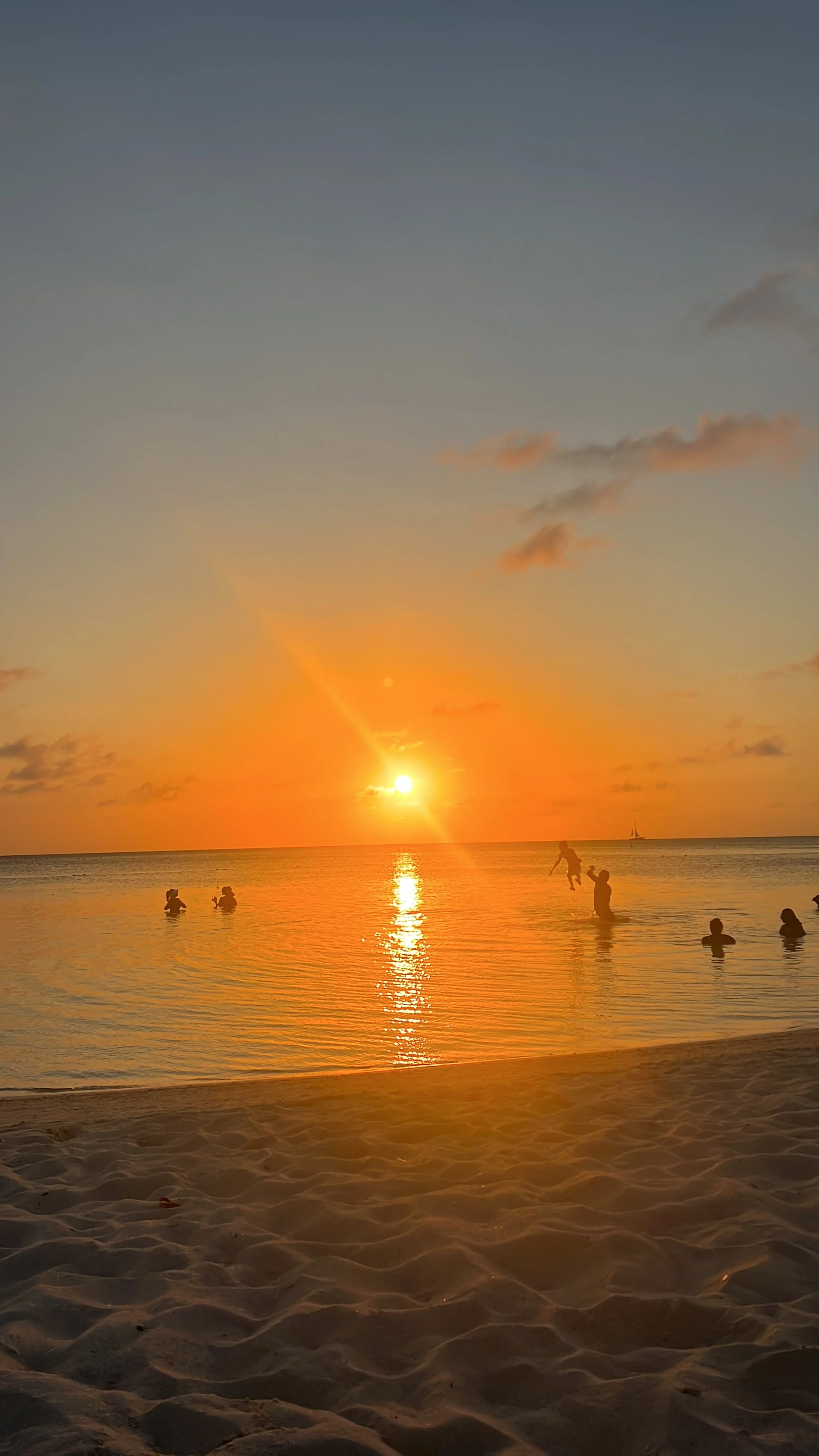 Beach scene at sunset with people swimming and playing in the water, sandy shore in the foreground, and a sailboat on the horizon.