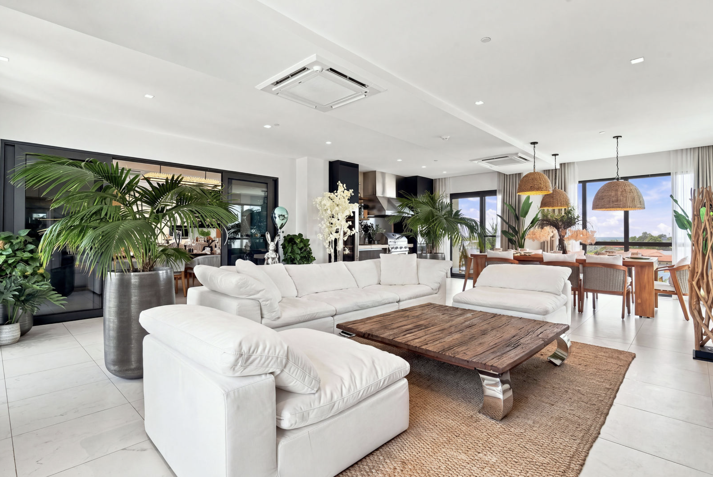 Modern open-concept living room with white sofas, indoor plants, a wooden coffee table, a dining area with woven pendant lights, and large windows showing a scenic view.