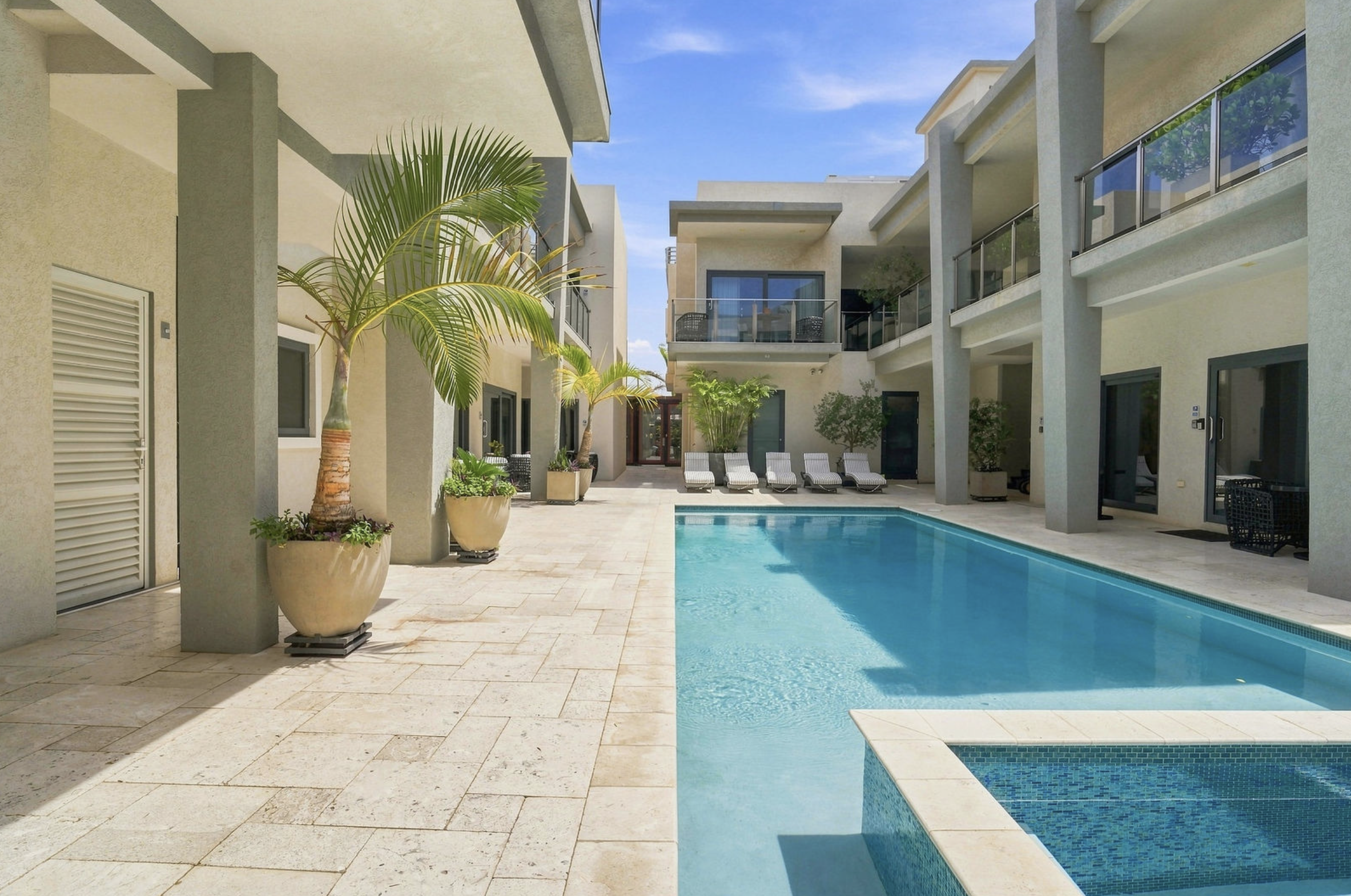 Modern apartment complex with poolside area, large potted palm trees, lounge chairs, and balconies, under a blue sky.