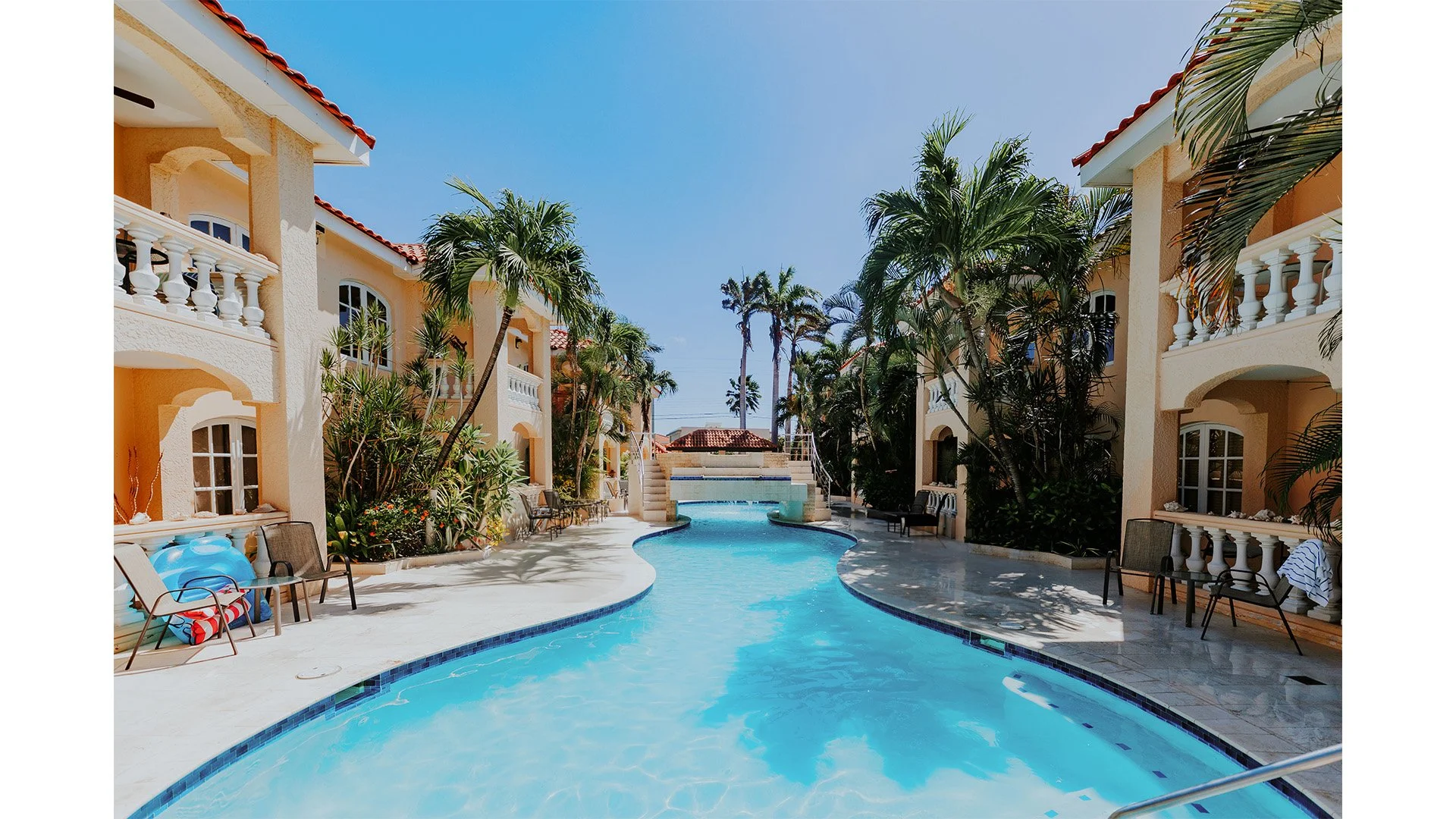 Poolside view of a vacation resort with palm trees, lounge chairs, and a small waterfall feature in the background.
