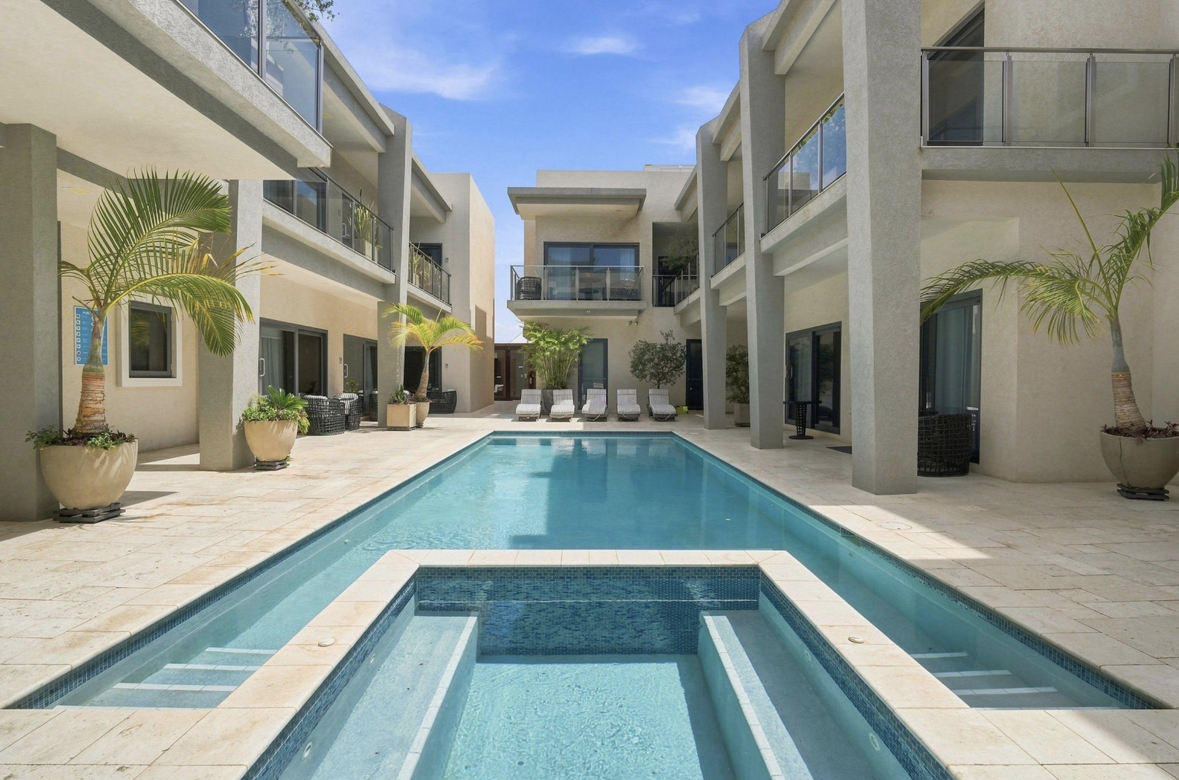 View of a modern apartment complex with a rectangular swimming pool and lounge chairs in the courtyard, surrounded by potted plants and balconies under a clear blue sky.