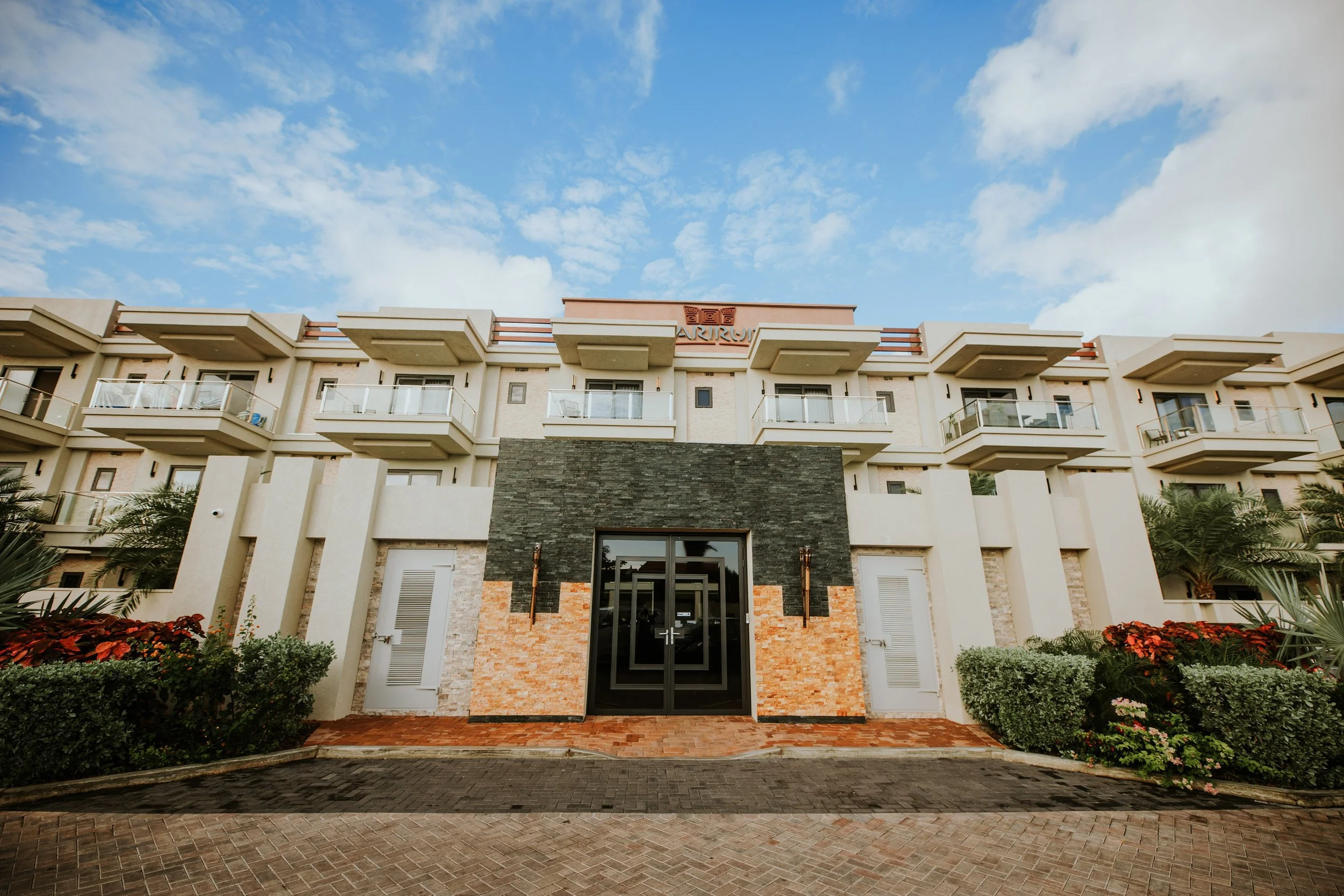 Front view of a modern multi-story apartment building with balconies, a black entrance door, and landscaping with bushes and flowers, under a cloudy blue sky.