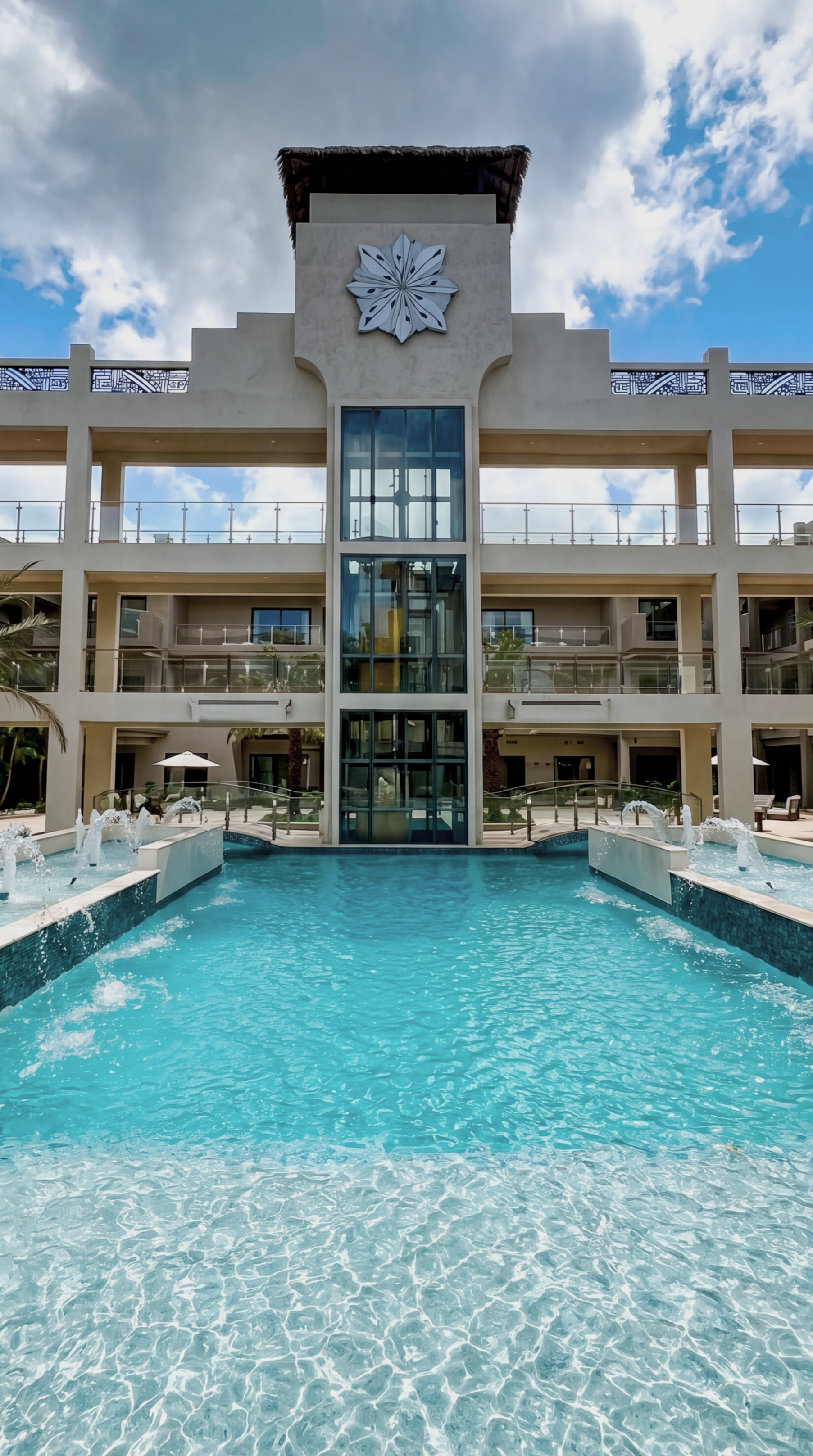 View of a hotel with a swimming pool in front, a glass elevator, and a decorative flower on the building facade. Blue sky with clouds in the background.