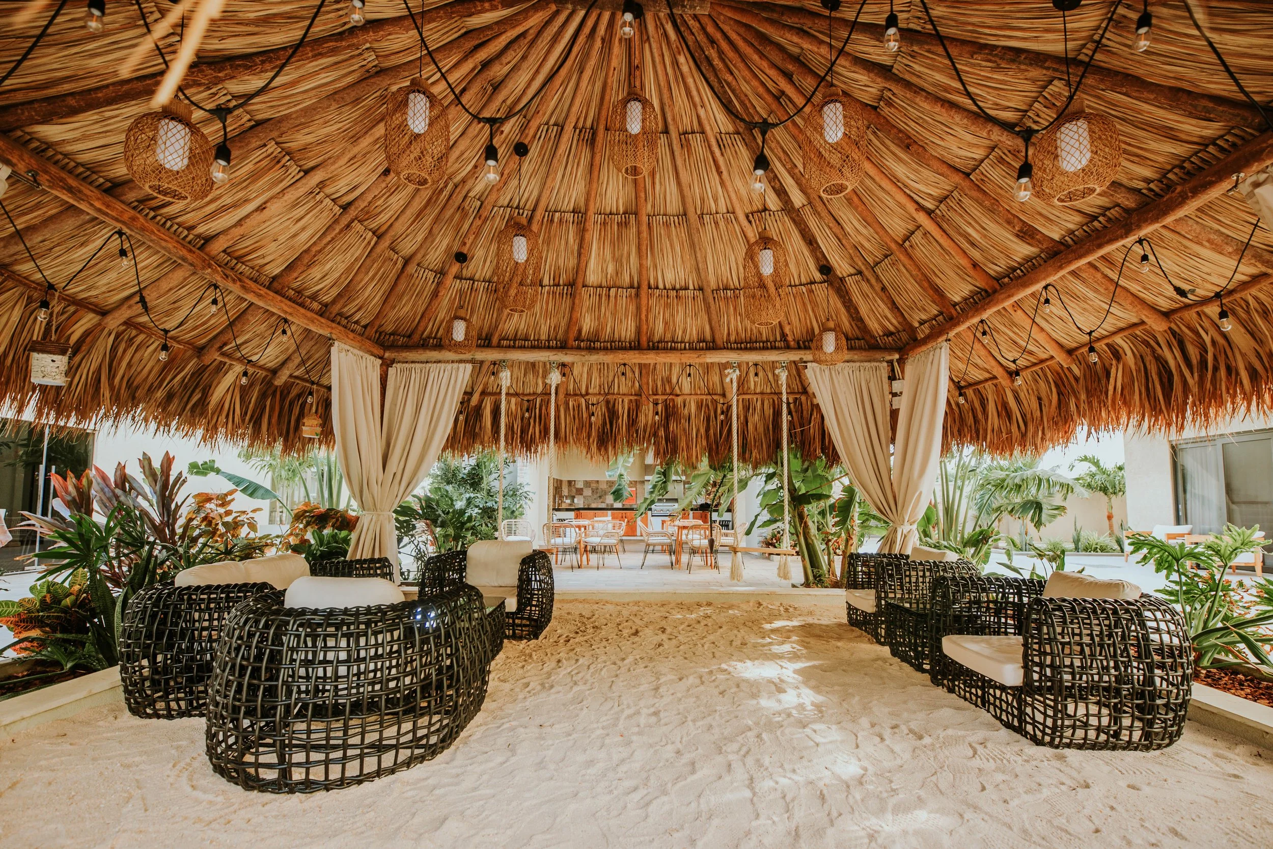 Outdoor seating area under a thatched roof with wicker chairs, cushioned seats, string lights, and surrounding tropical plants, with a sandy floor.