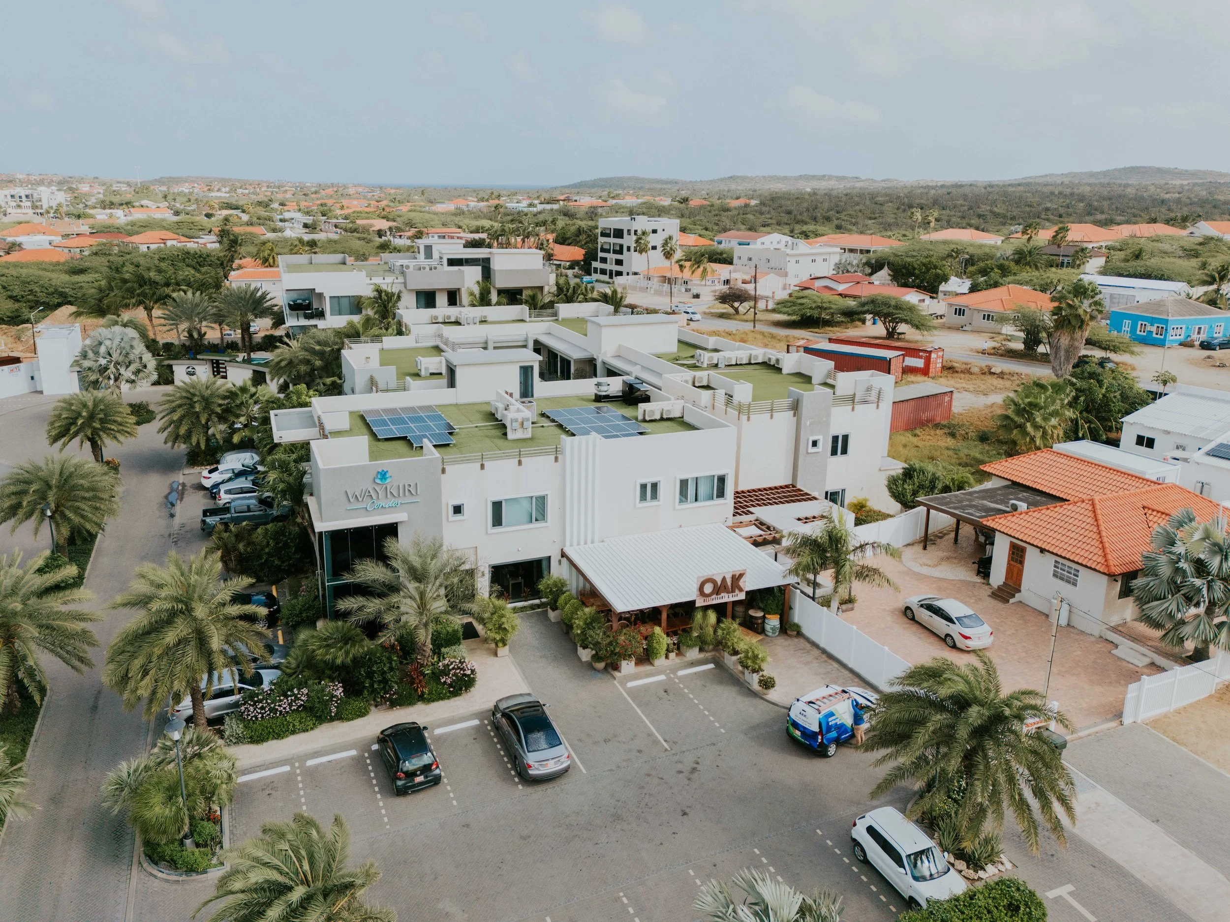 Aerial view of a modern resort with white buildings, solar panels on rooftops, surrounded by palm trees and parking lot with cars, located in a tropical area with other residential buildings and greenery in the background.