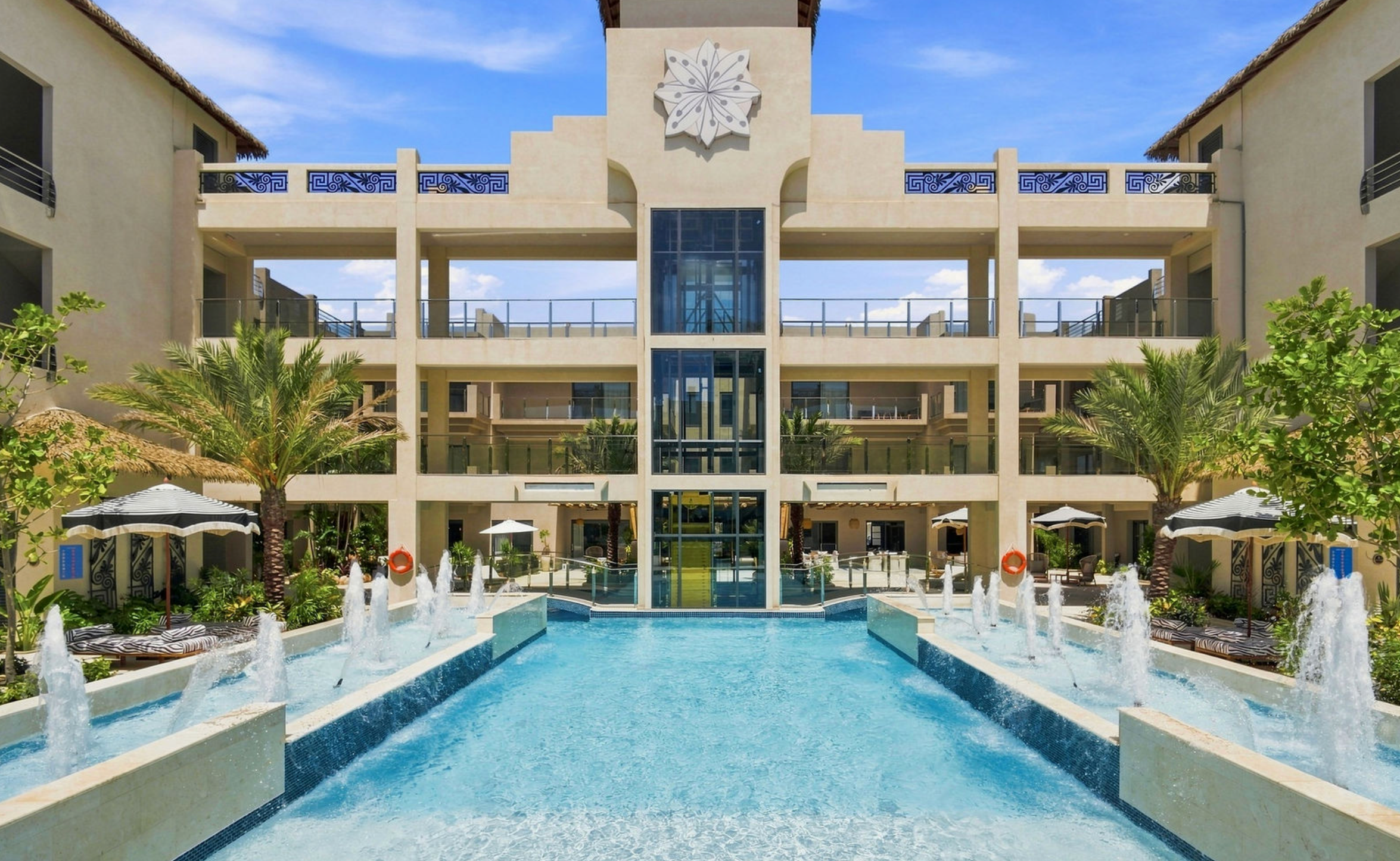 A modern hotel with a swimming pool, palm trees, and multiple balconies, under a blue sky with some clouds.
