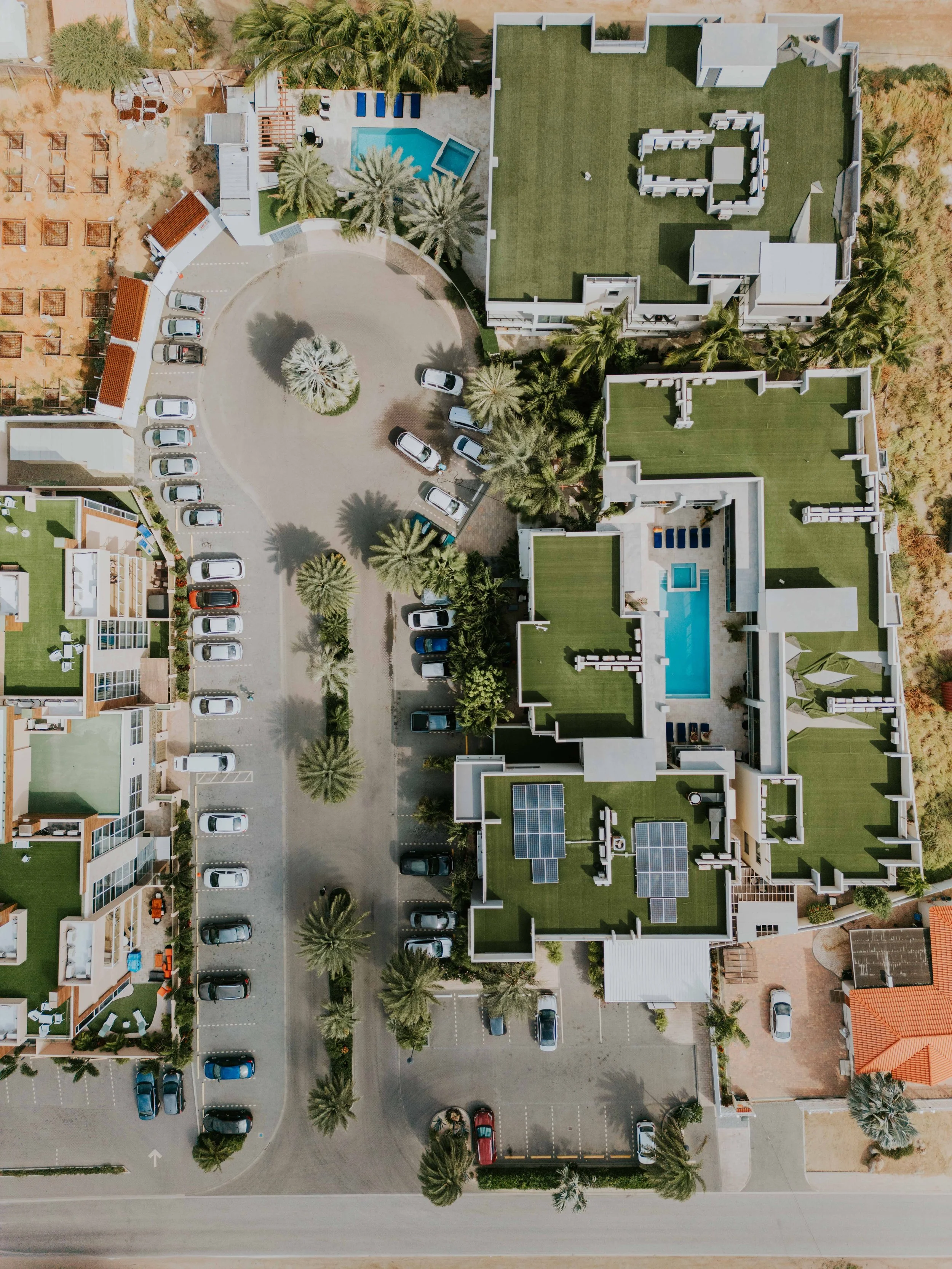 An aerial view of a residential complex with multiple buildings, green roofs, swimming pools, and parking lots surrounded by trees and a road.