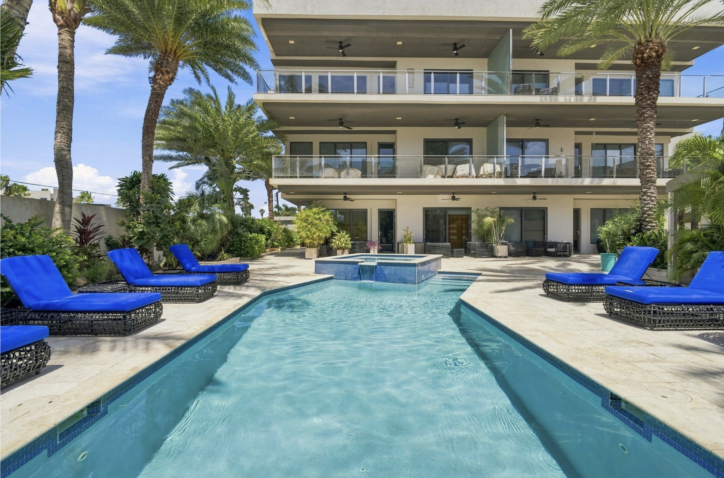 Luxury apartment building with balconies, overlooking a backyard with a swimming pool surrounded by blue lounge chairs and lush tropical plants under a clear blue sky.