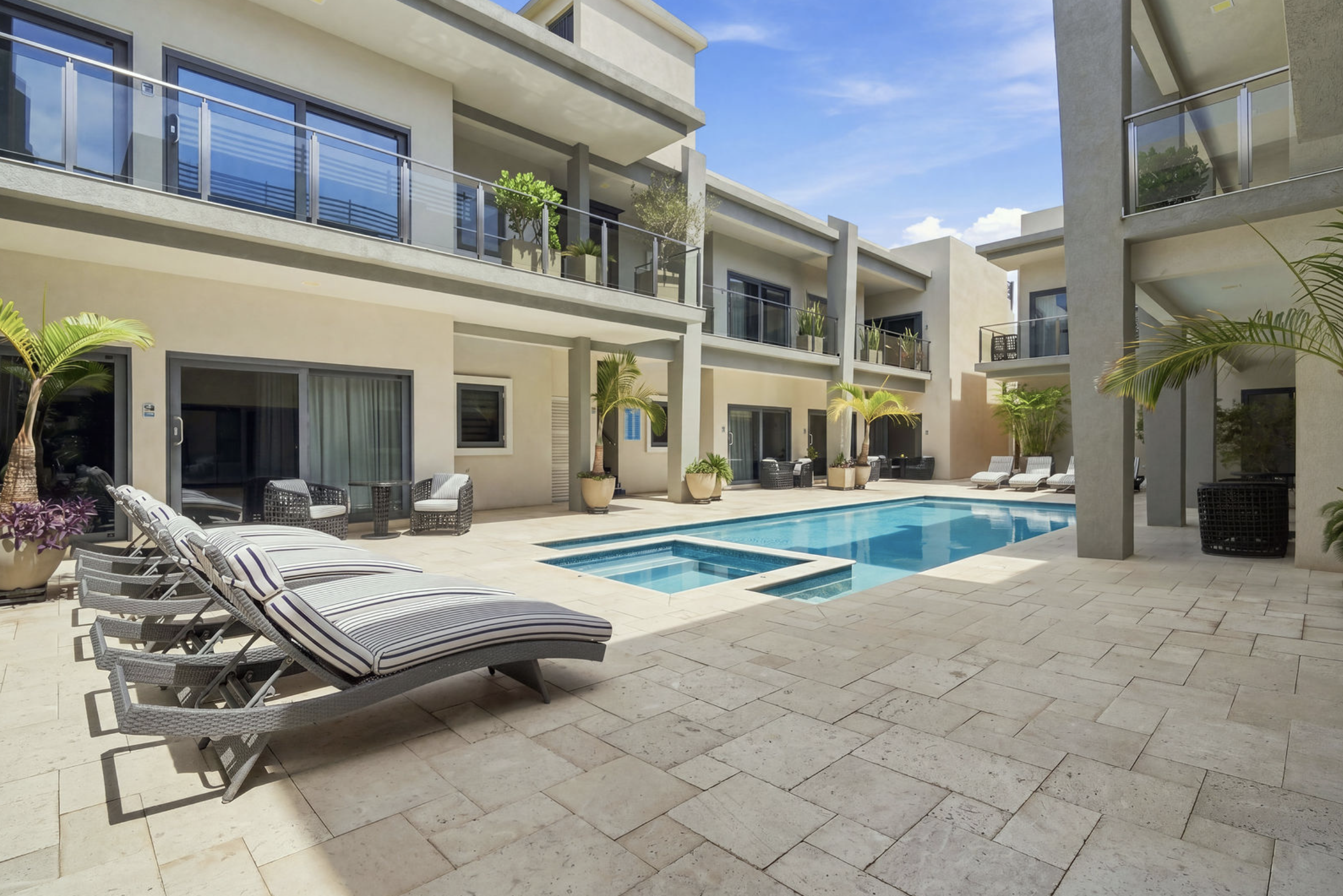 View of a modern residential complex with a swimming pool, lounge chairs, and balcony areas with railings and plants, under a clear blue sky.