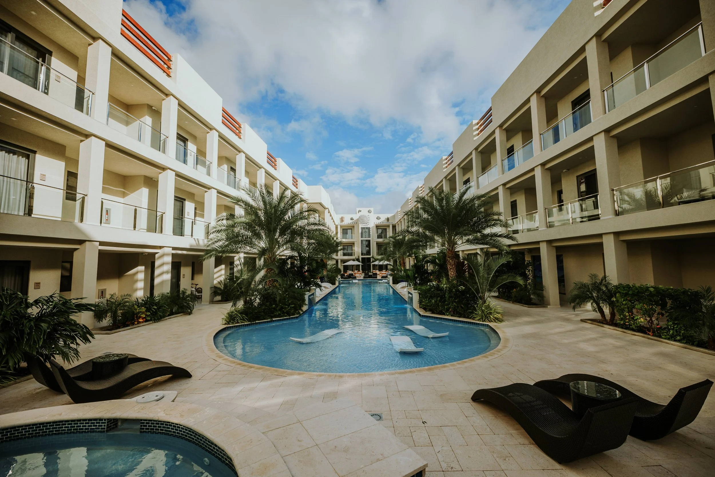 View of a hotel courtyard with a swimming pool flanked by lounge chairs and tropical plants, surrounded by modern, multi-story hotel buildings with balconies.