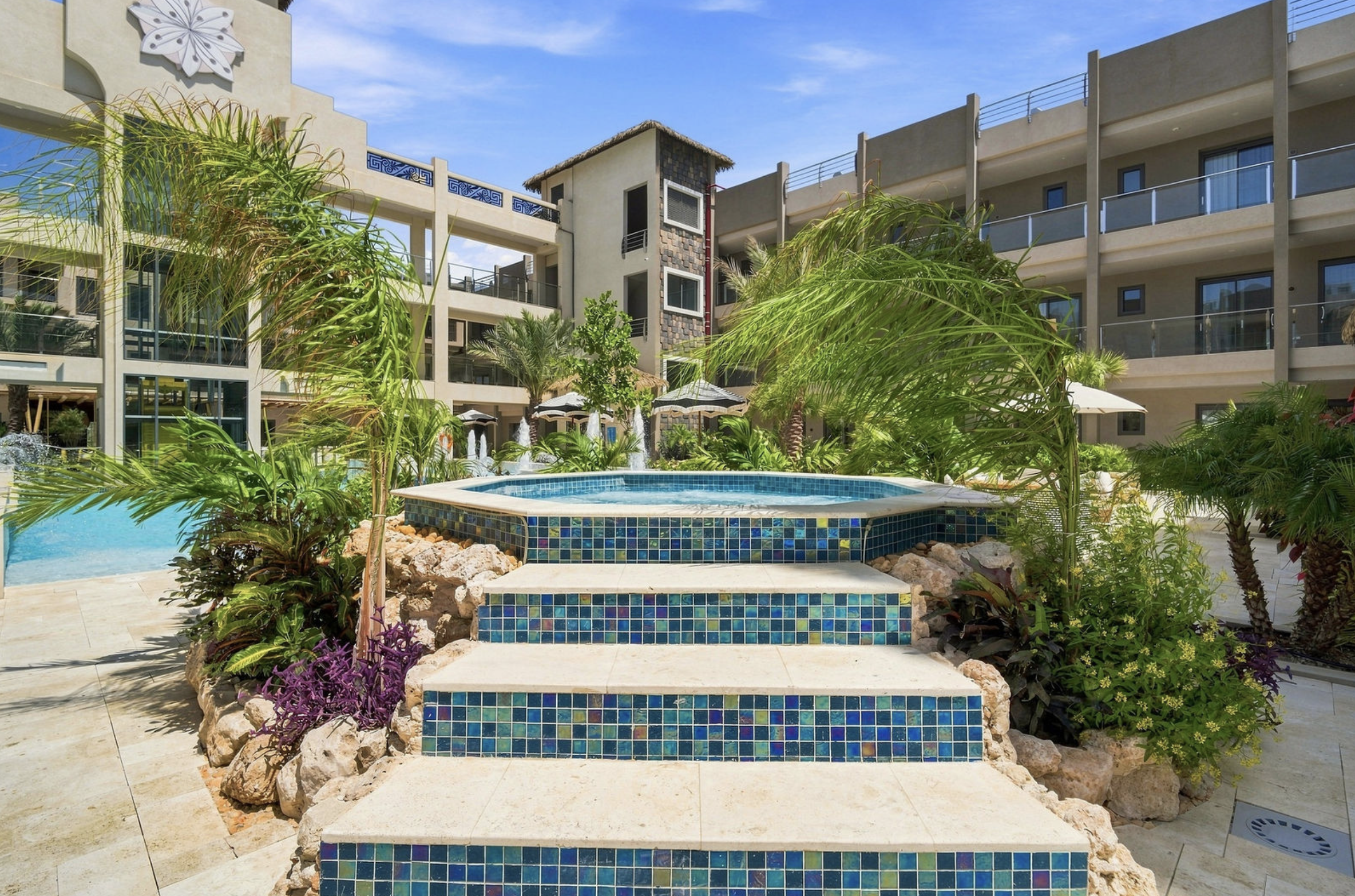 A swimming pool area in a modern hotel courtyard surrounded by lush tropical plants, with multi-story hotel buildings in the background and a bright blue sky overhead.