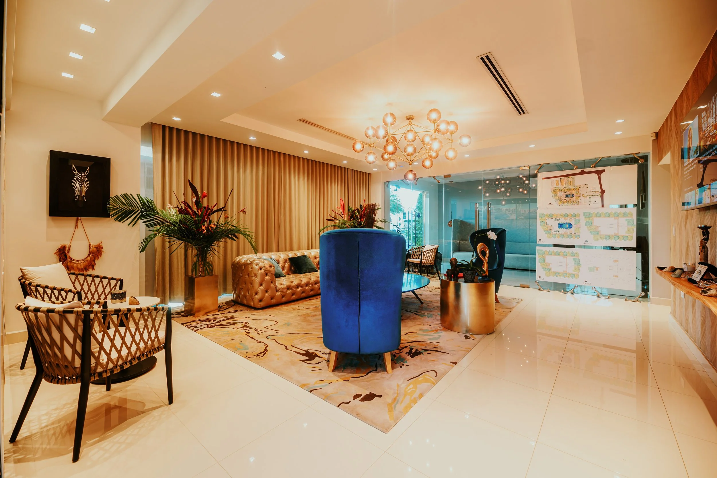 Elegant hotel lobby with modern decor, featuring a beige tufted sofa, a blue velvet armchair, gold side tables, large floral arrangements, and a contemporary chandelier. There are seating areas, wall art, and a glass wall with floor plans.