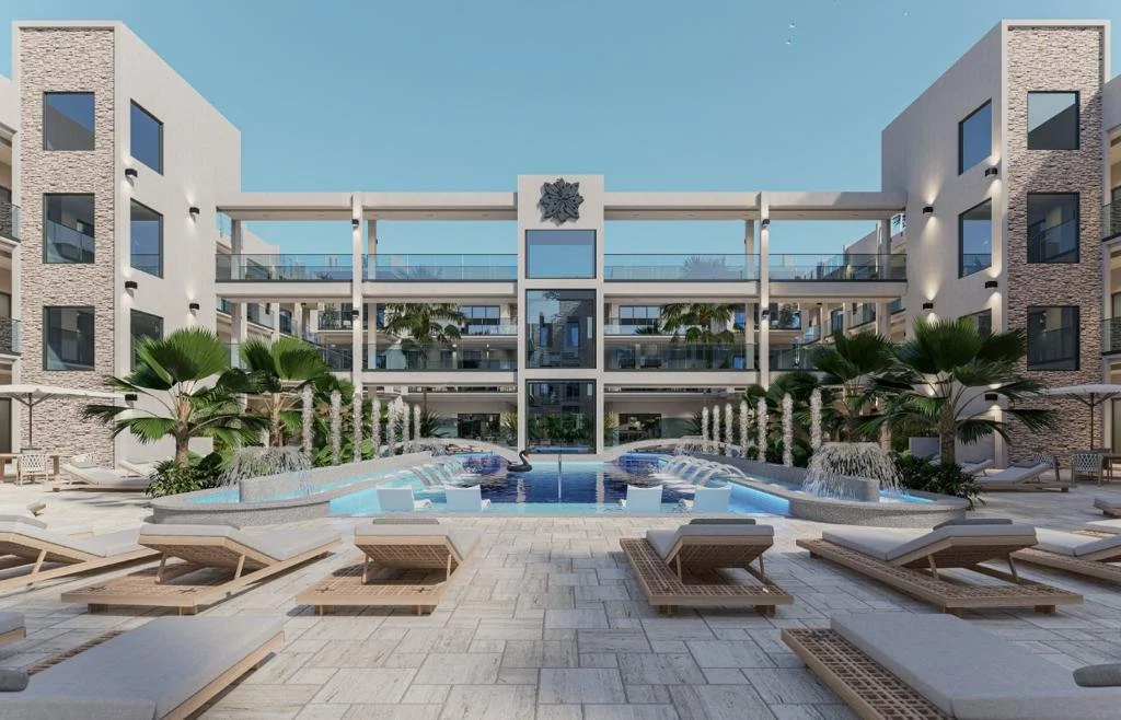 Modern apartment complex courtyard with a swimming pool, palm trees, lounge chairs, fountains, and a building with balconies and glass railings.