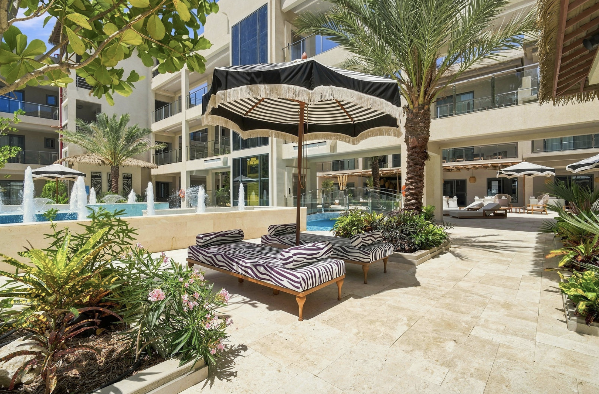 A luxurious hotel pool area with lounge chairs featuring zebra print cushions, shaded by black and white striped umbrellas. Tall palm trees and lush green plants surround the space, with a modern multi-story building in the background. There are wate