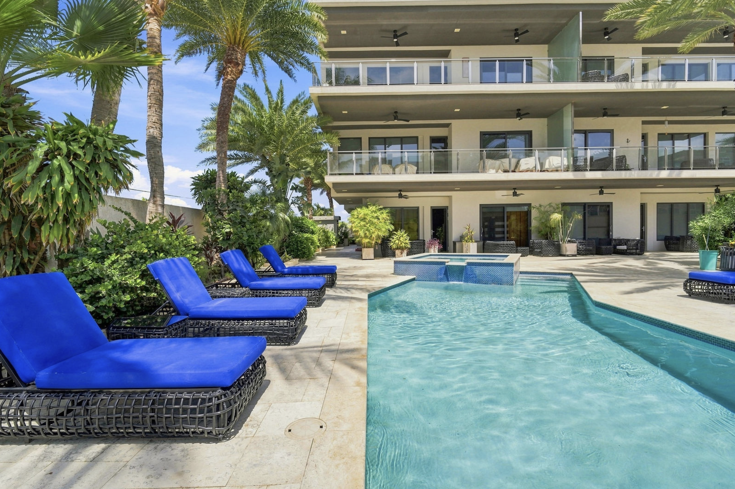 A modern apartment building with balconies overlooking a backyard with a pool, surrounded by lush tropical plants and palm trees, under a blue sky.