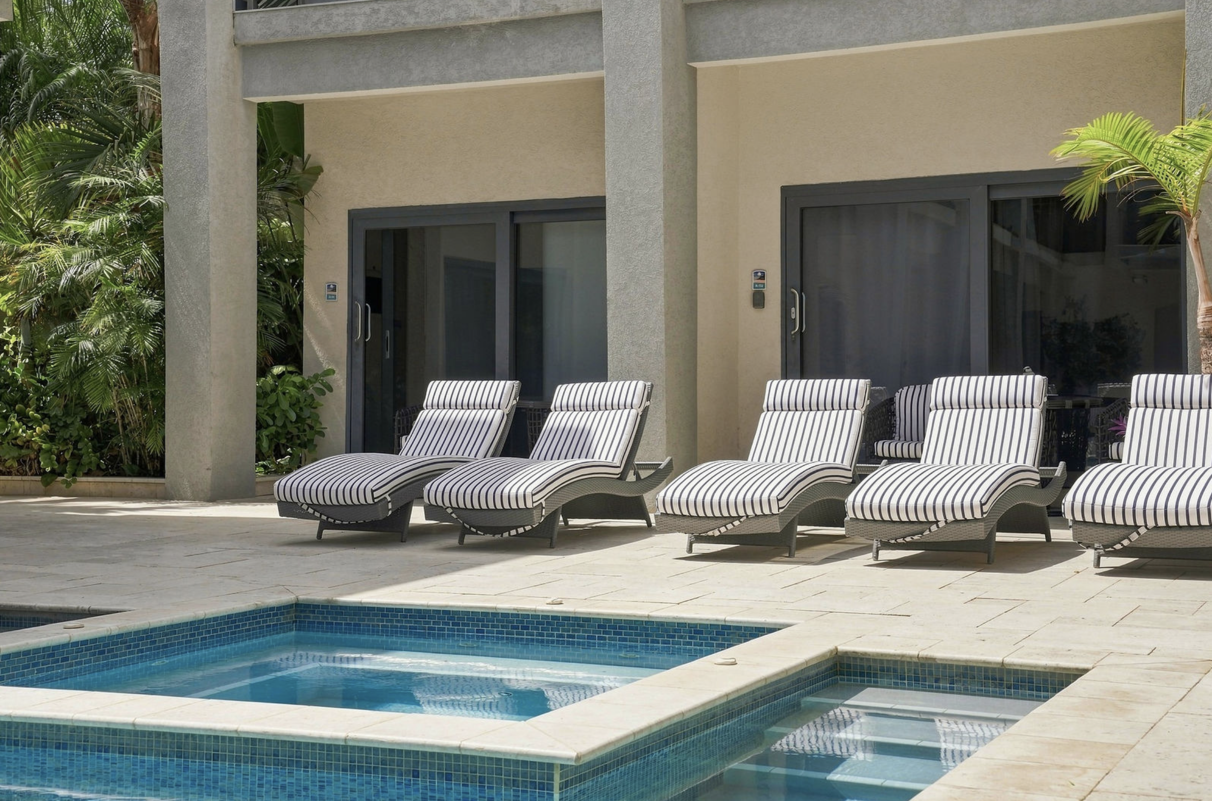 Pool area with lounge chairs, a swimming pool, and a building with sliding glass doors and embedded security keypads, surrounded by tropical plants.