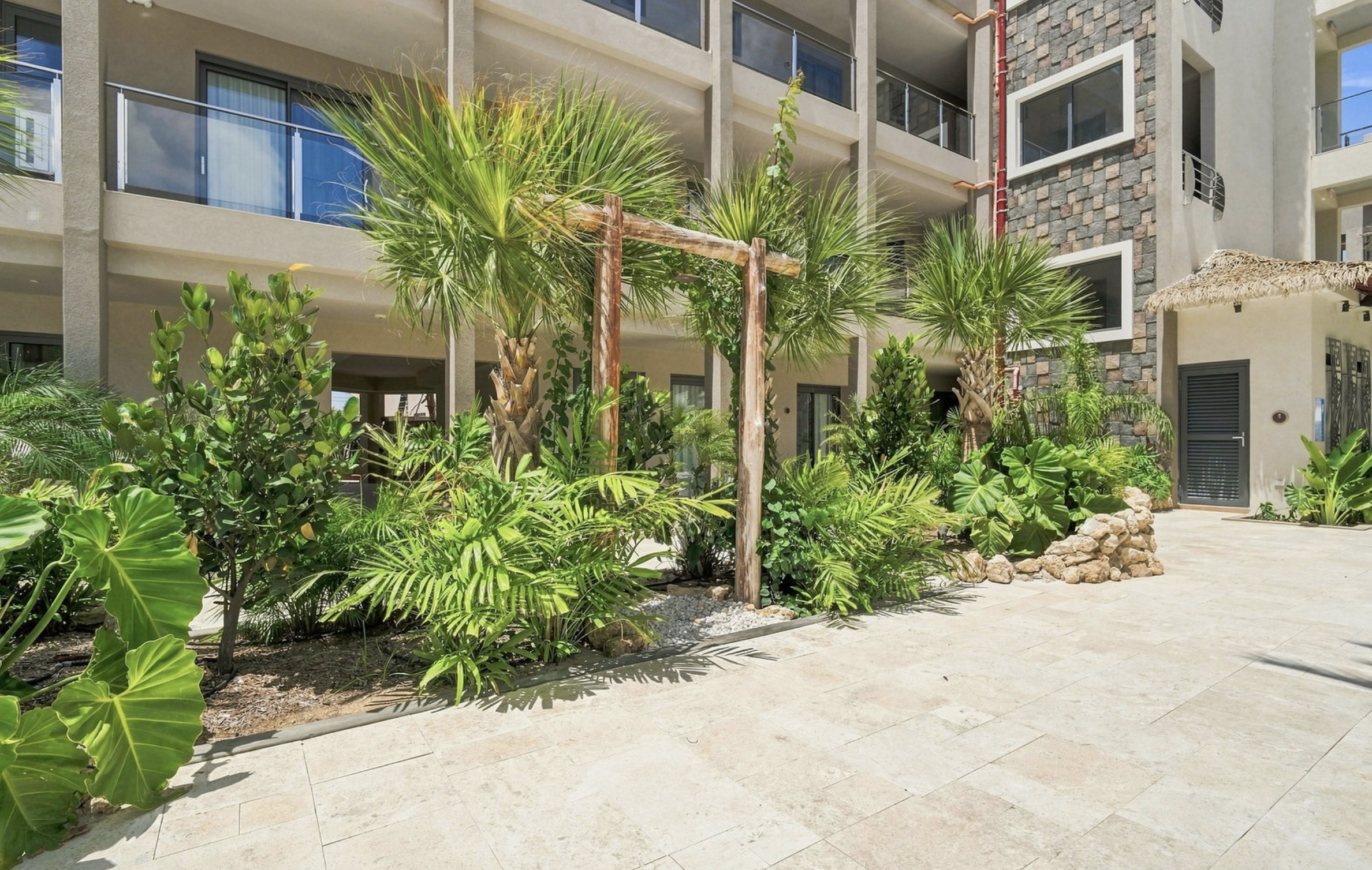 A view of an outdoor patio area in front of a modern apartment building, featuring tropical plants, palm trees, and a stone pathway with a thatched umbrella in the background.
