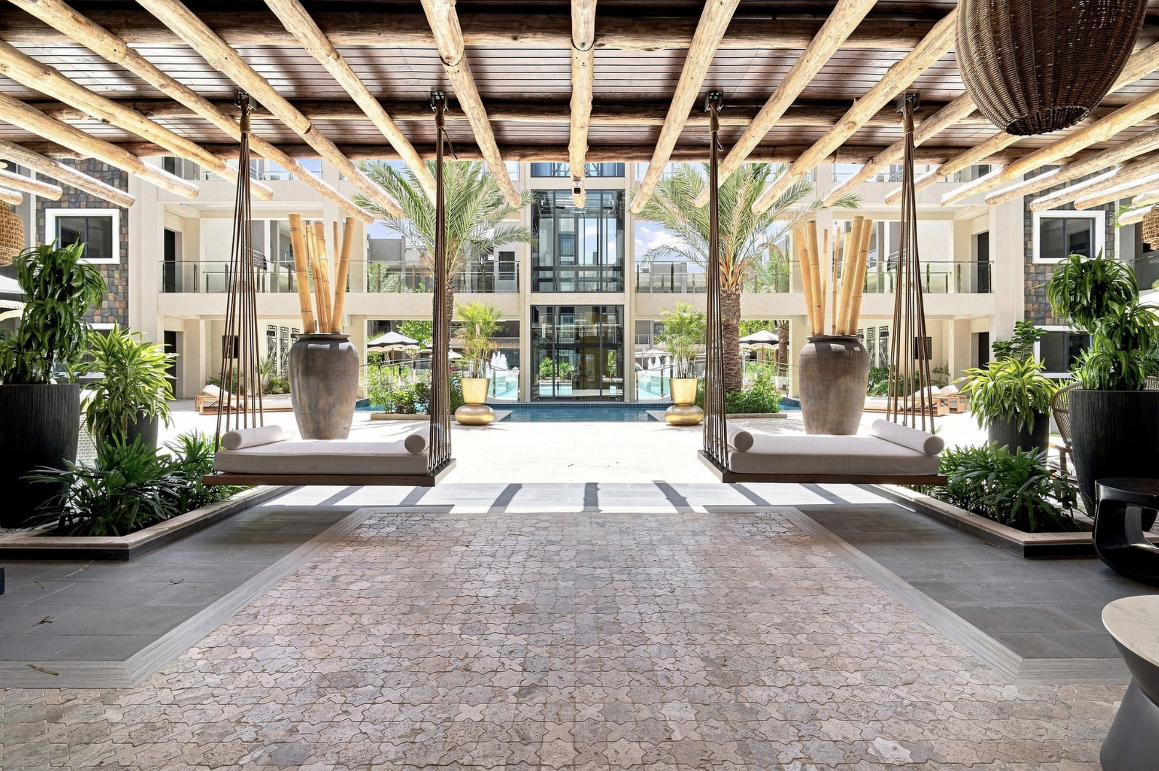 A covered outdoor lounge area with two swing chairs, potted plants, and a view of a swimming pool and modern apartment building in the background.