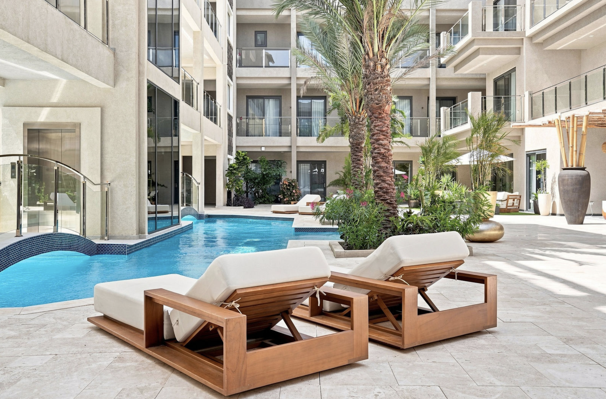 Outdoor swimming pool with lounge chairs, a large potted plant, and a tall palm tree surrounded by apartment or hotel building with balconies, glass railings, and sliding glass doors.