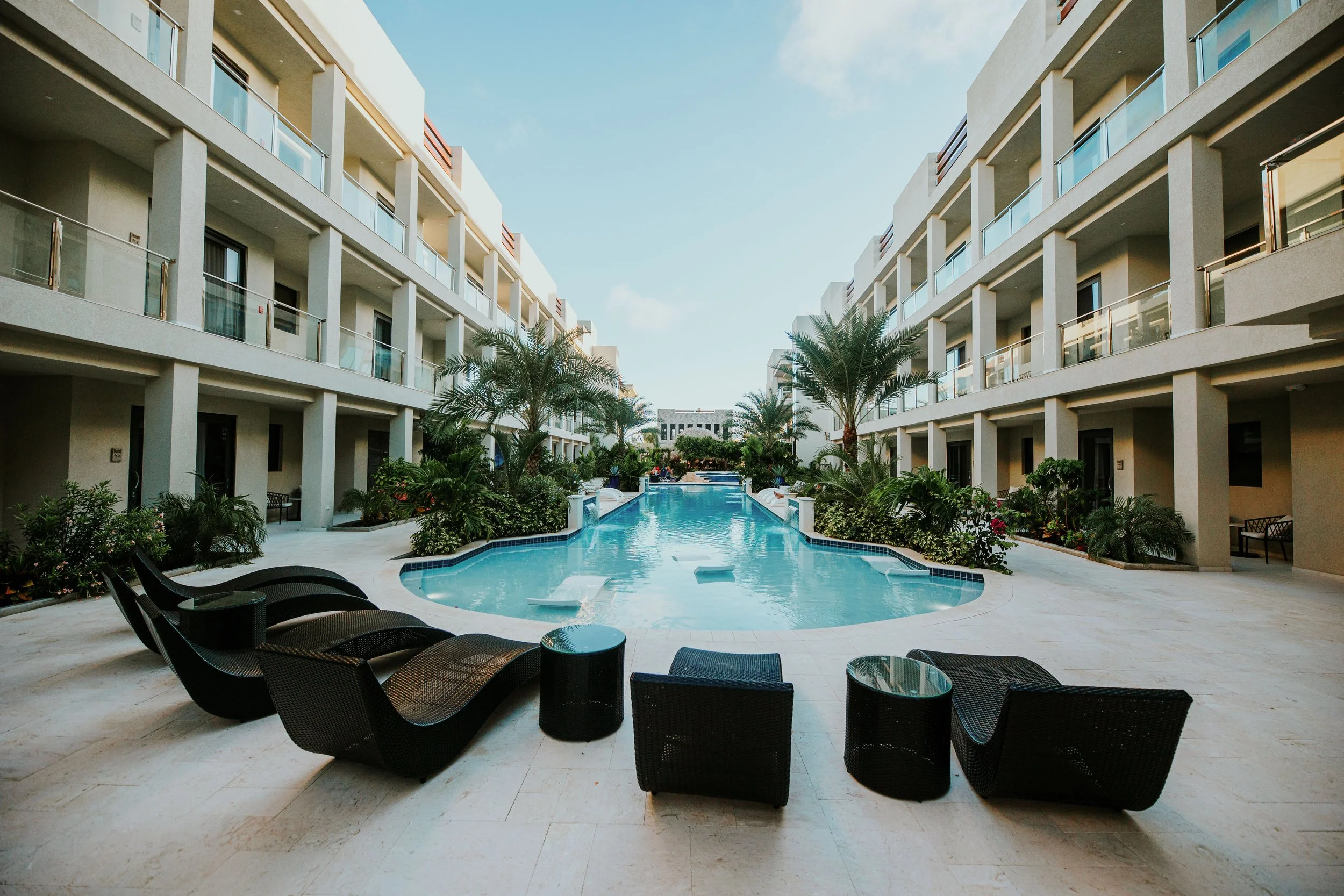 A modern hotel courtyard with a swimming pool, lounge chairs, and tropical plants, surrounded by multi-story buildings with balconies.