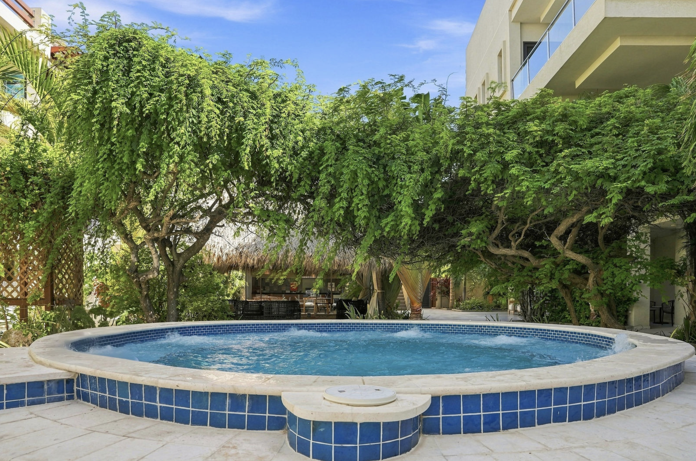 A backyard scene featuring a circular swimming pool with blue tile edging, surrounded by white stone paving. In the background, tall green trees and a patio area with outdoor furniture and a thatched-roof cabana are visible, along with a modern multi