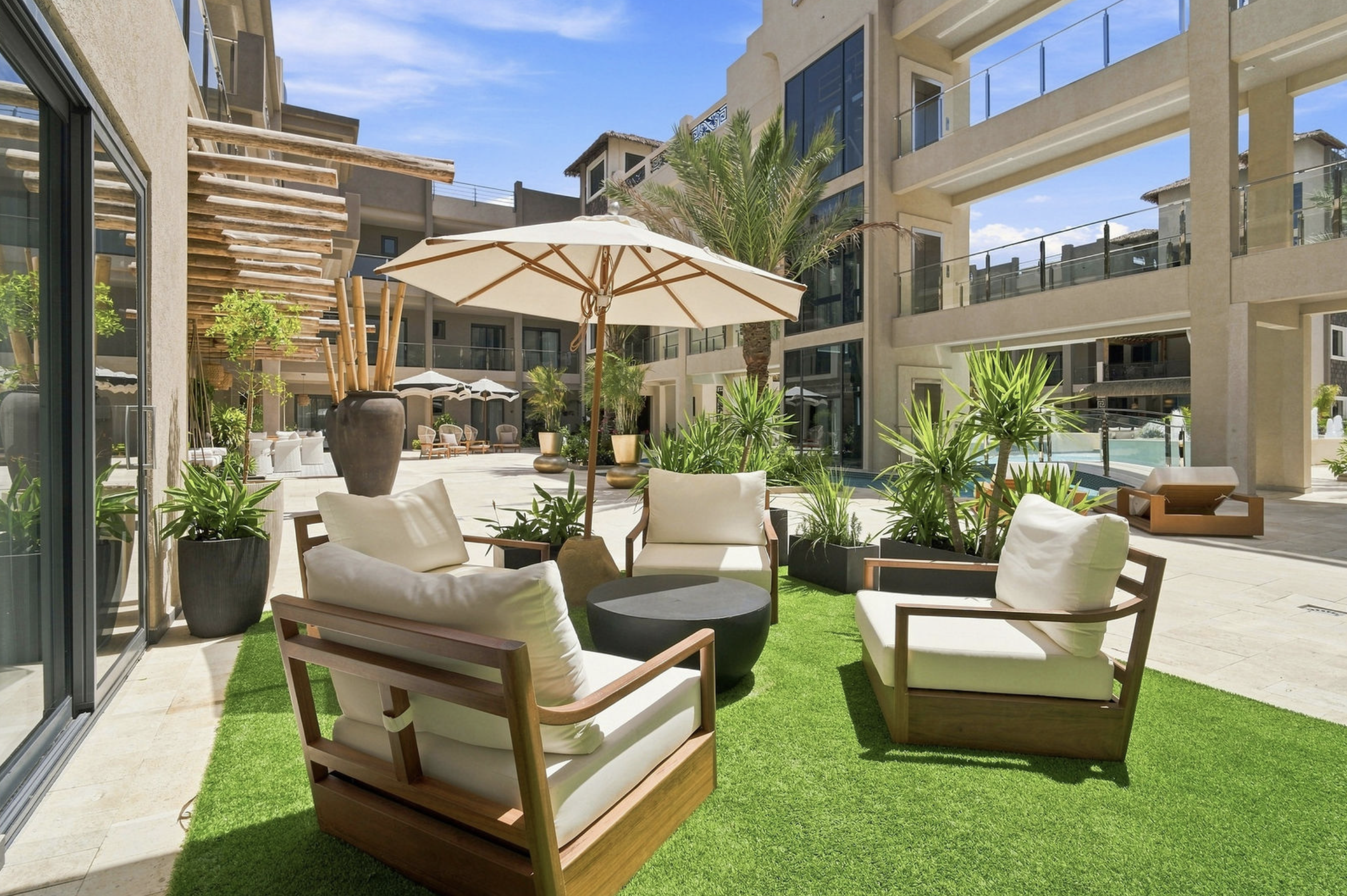 An outdoor seating area with white cushioned chairs, small tables, and green artificial grass, surrounded by potted plants, and shaded by umbrellas, with modern multi-story buildings and a pool in the background.