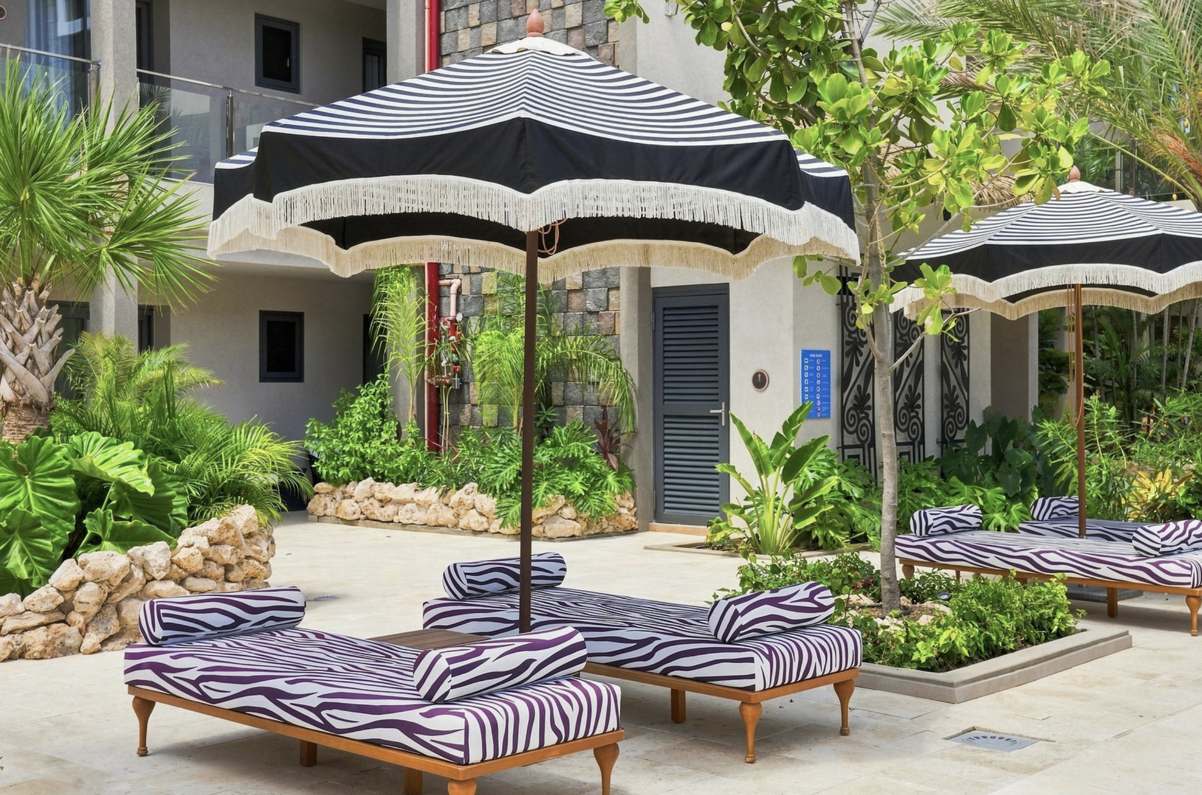 Poolside area with zebra-striped lounge chairs, black and white striped umbrellas, lush green plants, and a modern building in the background.