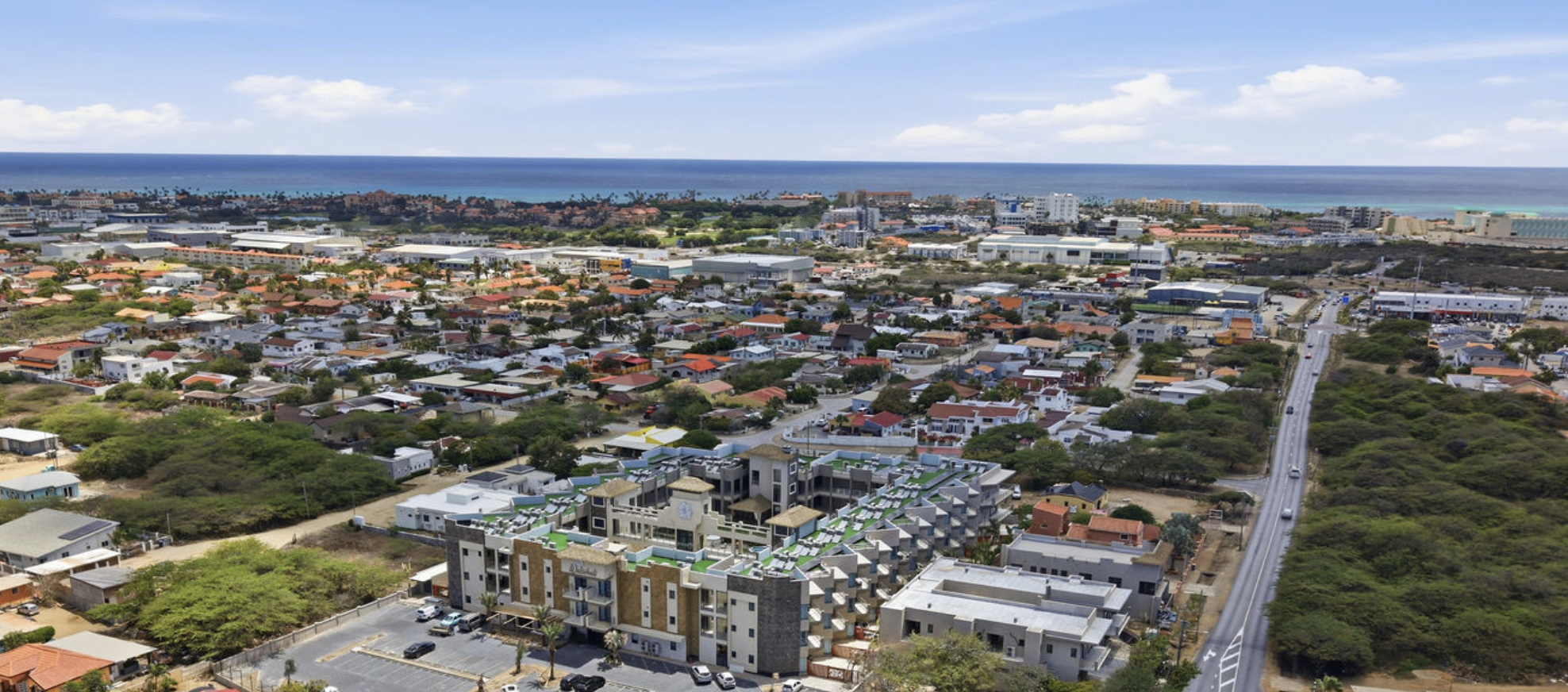 Aerial view of a coastal city with residential neighborhoods, commercial buildings, and a road leading toward the ocean in the background under partly cloudy skies.