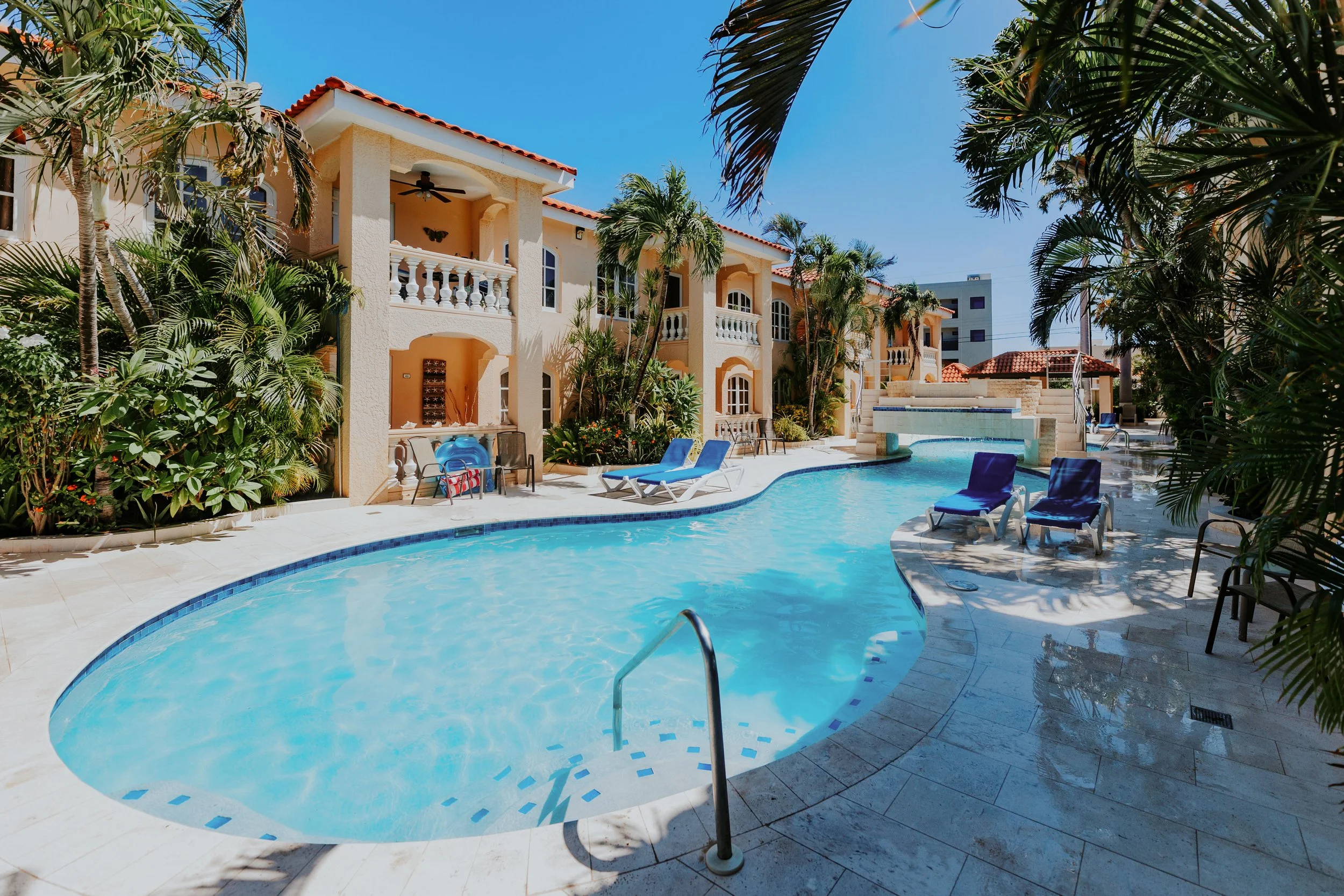 A resort swimming pool area surrounded by palm trees and pastel-colored buildings with balconies, under a clear blue sky.
