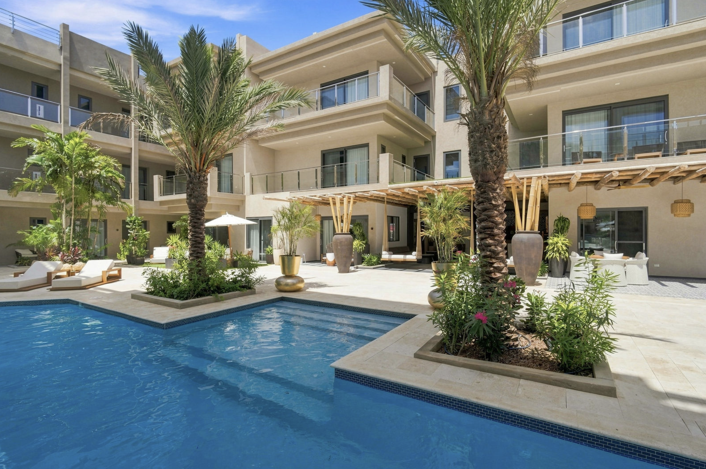 Luxury apartment complex with a swimming pool surrounded by palm trees, chairs, and lush greenery under a sunny sky.