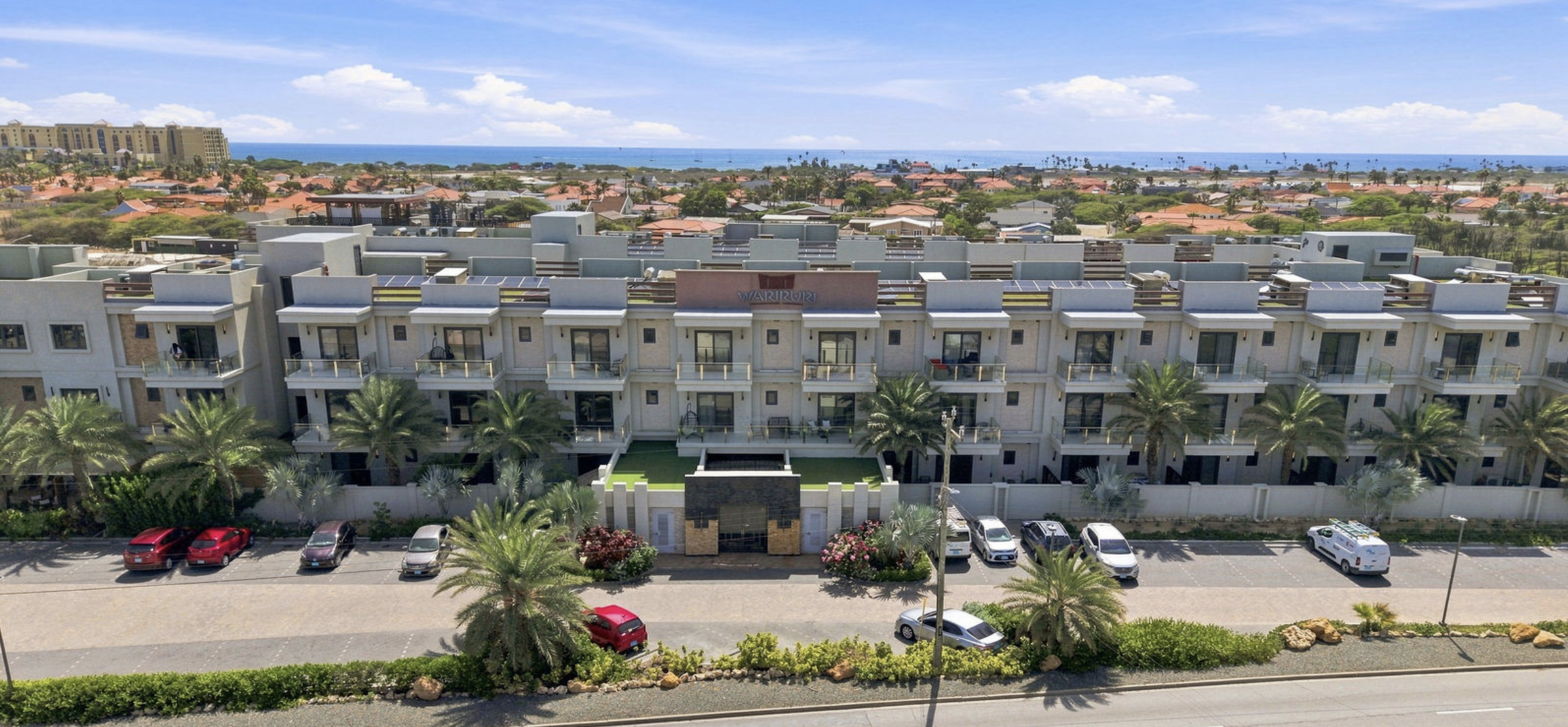 An aerial view of a modern beachfront hotel with balconies, palm trees, parked cars in front, and a beach and ocean in the background.