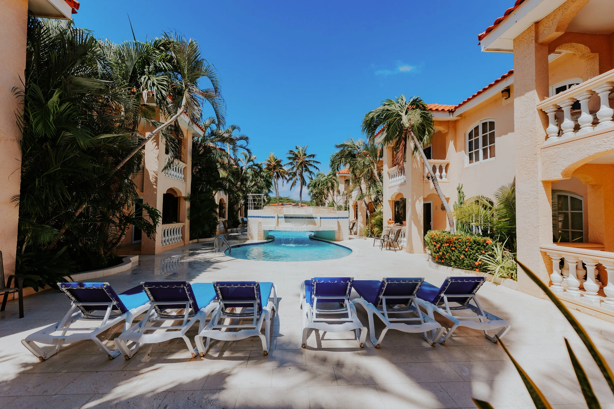 Poolside area at a tropical resort, featuring blue lounge chairs, palm trees, a swimming pool, and a waterfall feature in the background, surrounded by peach-colored buildings with balconies under a clear blue sky.