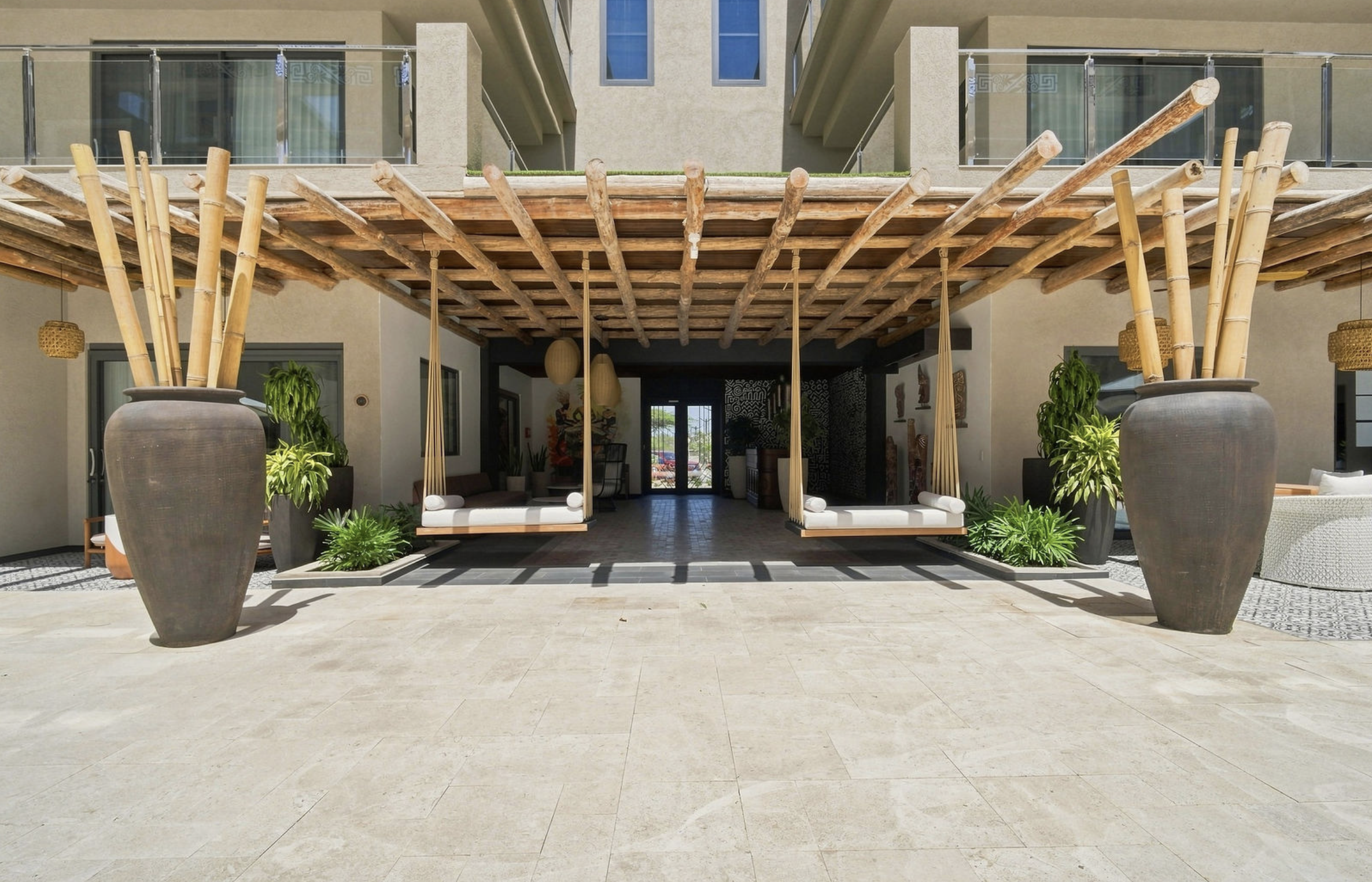 Outdoor patio with two large pots holding tall bamboo plants, two hanging swing chairs, and potted plants, with a building in the background.