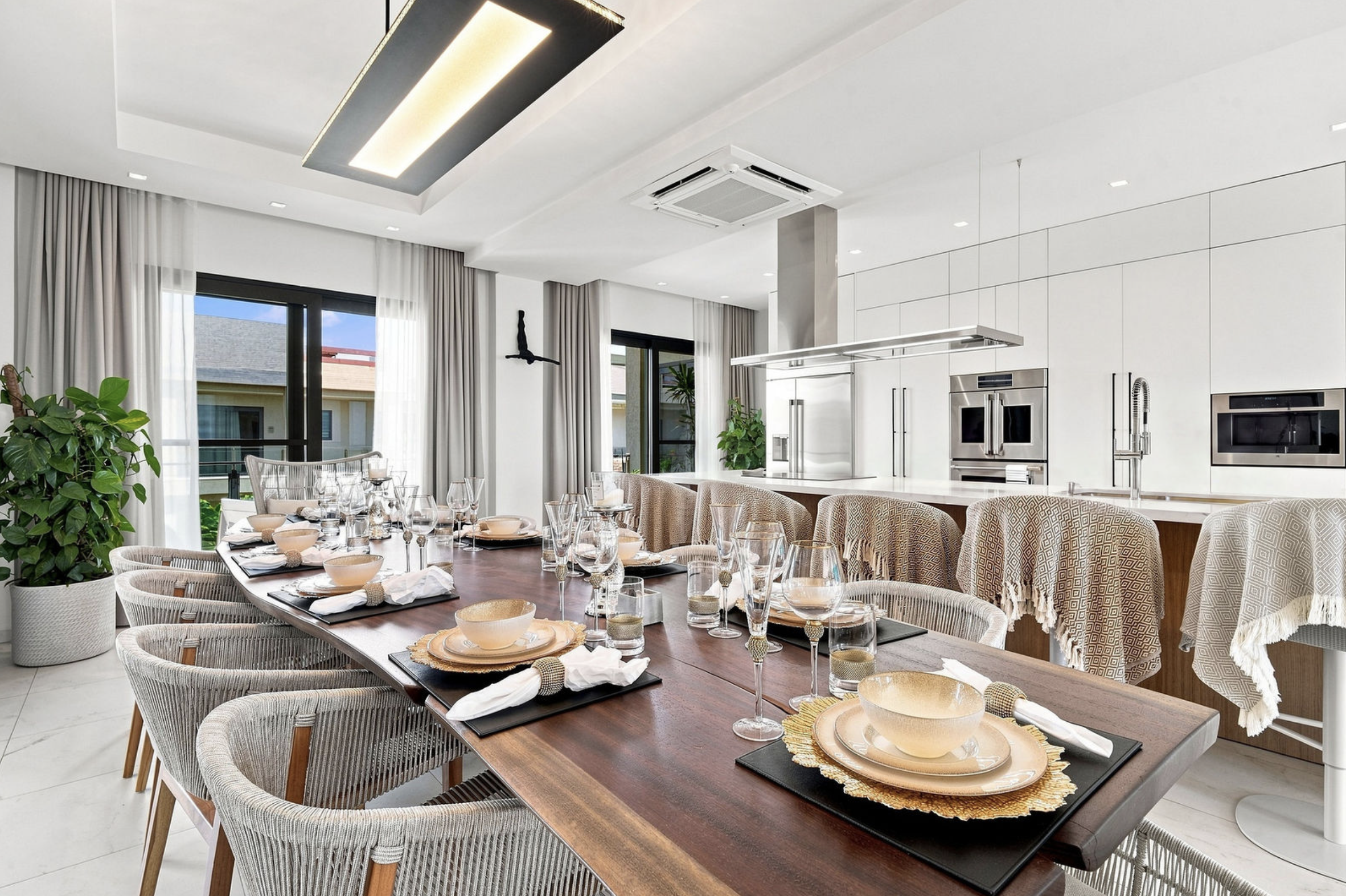 Modern open-concept kitchen and dining area with a large wooden table set with dinnerware, glassware, and place settings. The space features white walls, large windows with white curtains, and a kitchen with built-in stainless steel appliances and wh